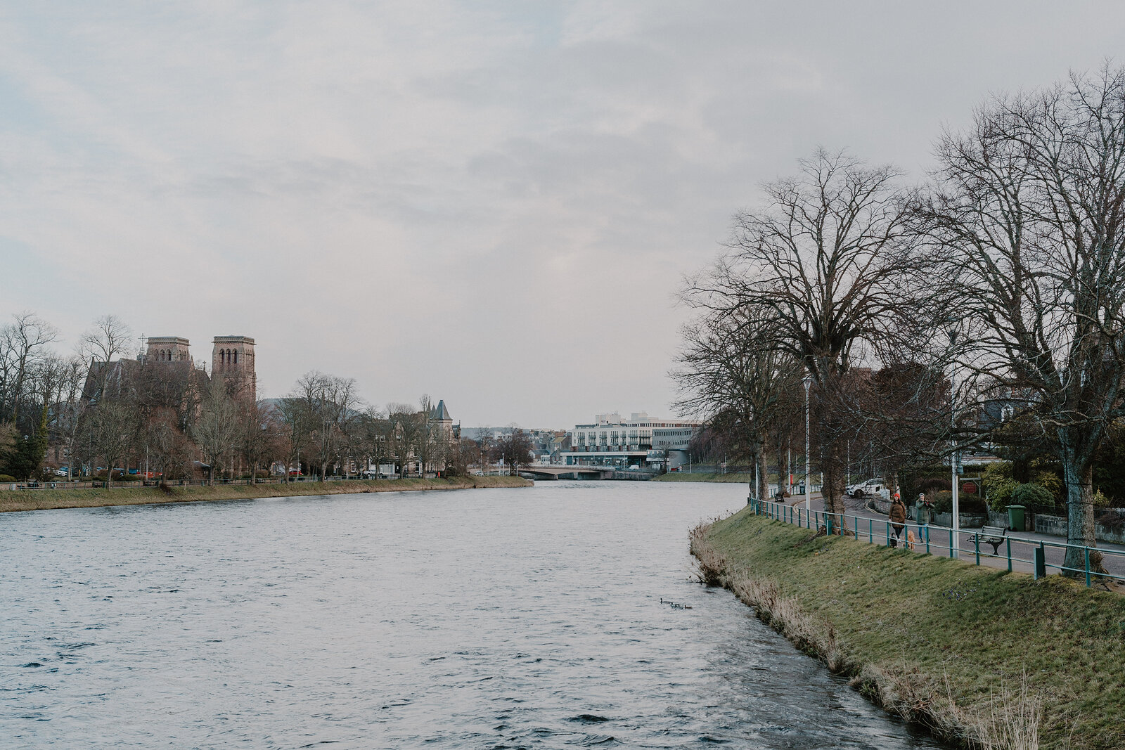 A calm river winds past a park lined with bare trees toward twin church towers on the far bank beneath an overcast sky.