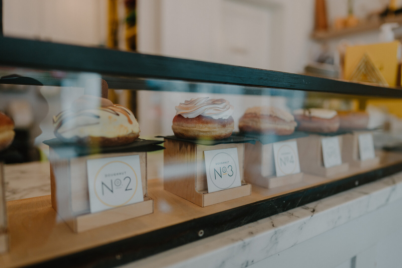 A glass display case holds four glazed doughnuts each labelled with a numbered card, marble counter visible below.