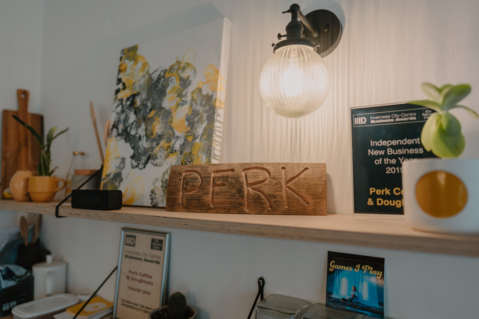 A carved wooden sign reading 'PERK' sits on a shelf beside a framed abstract painting, an award certificate and potted succulents.