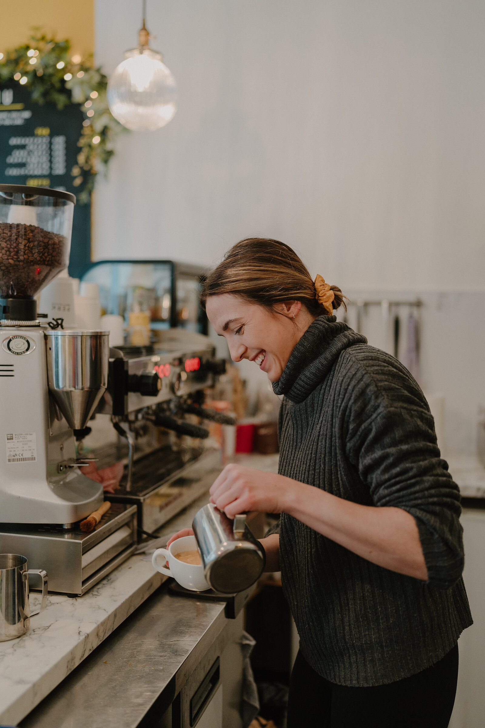 A smiling woman steams milk at an espresso machine, a coffee bean hopper and chalkboard menu visible behind her.