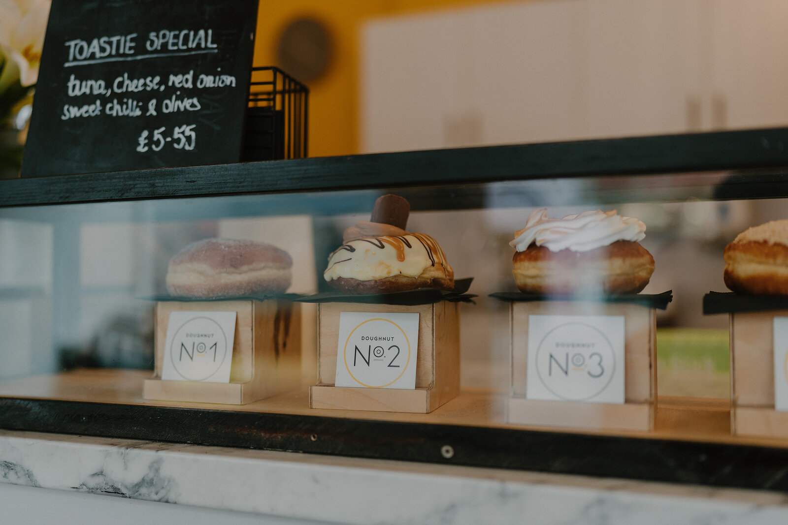A glass display case holds three glazed doughnuts numbered 1 to 3 beside a chalkboard advertising a toastie special.