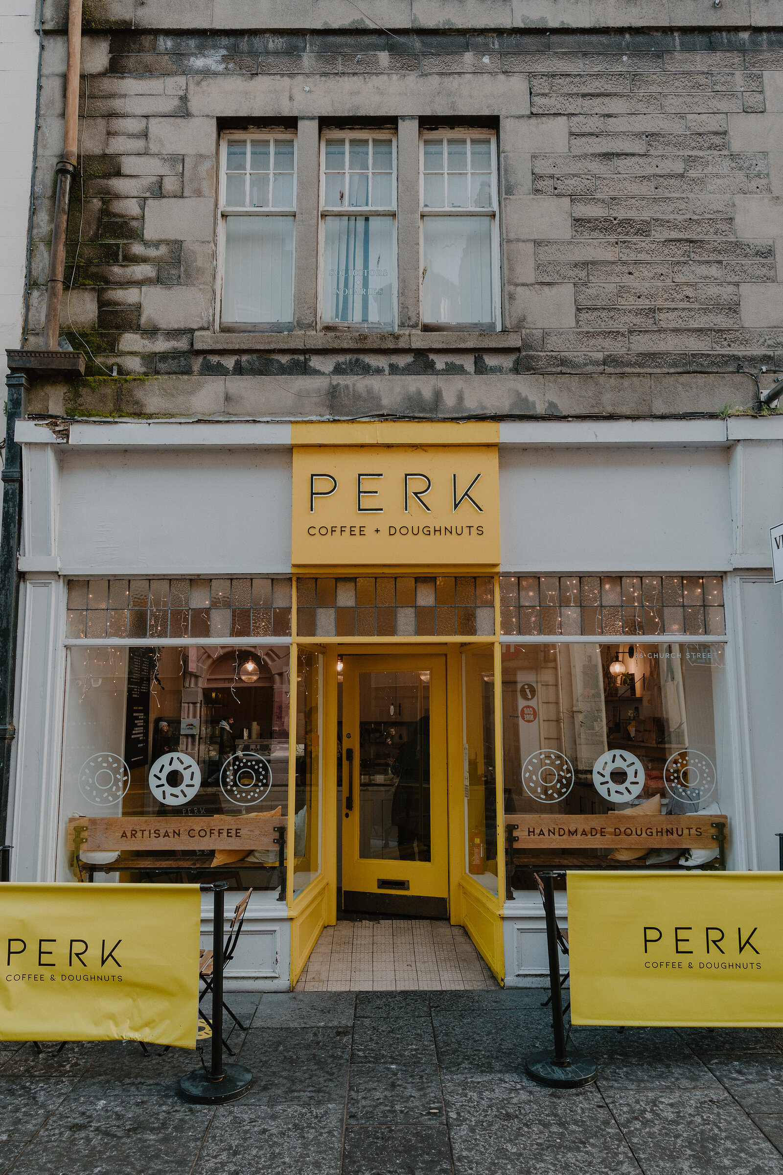 A yellow shopfront carries a sign reading 'Perk, Coffee + Doughnuts' above a lit entrance and window displays.