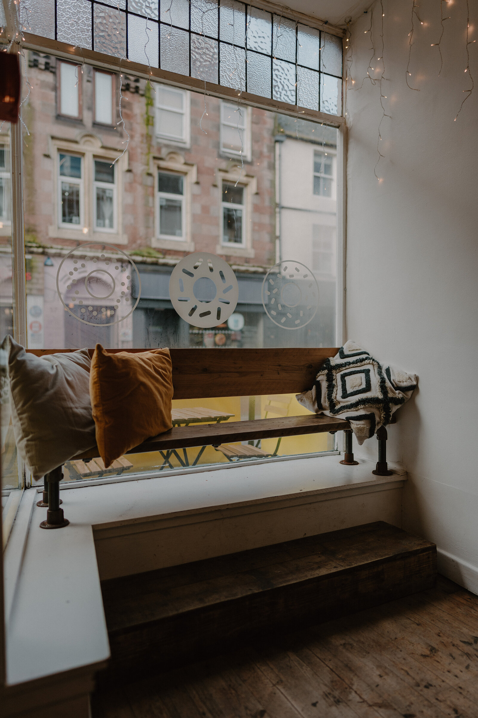 A wooden bench with patterned cushions sits beneath a frosted window etched with doughnut shapes, string lights draped along the glass.