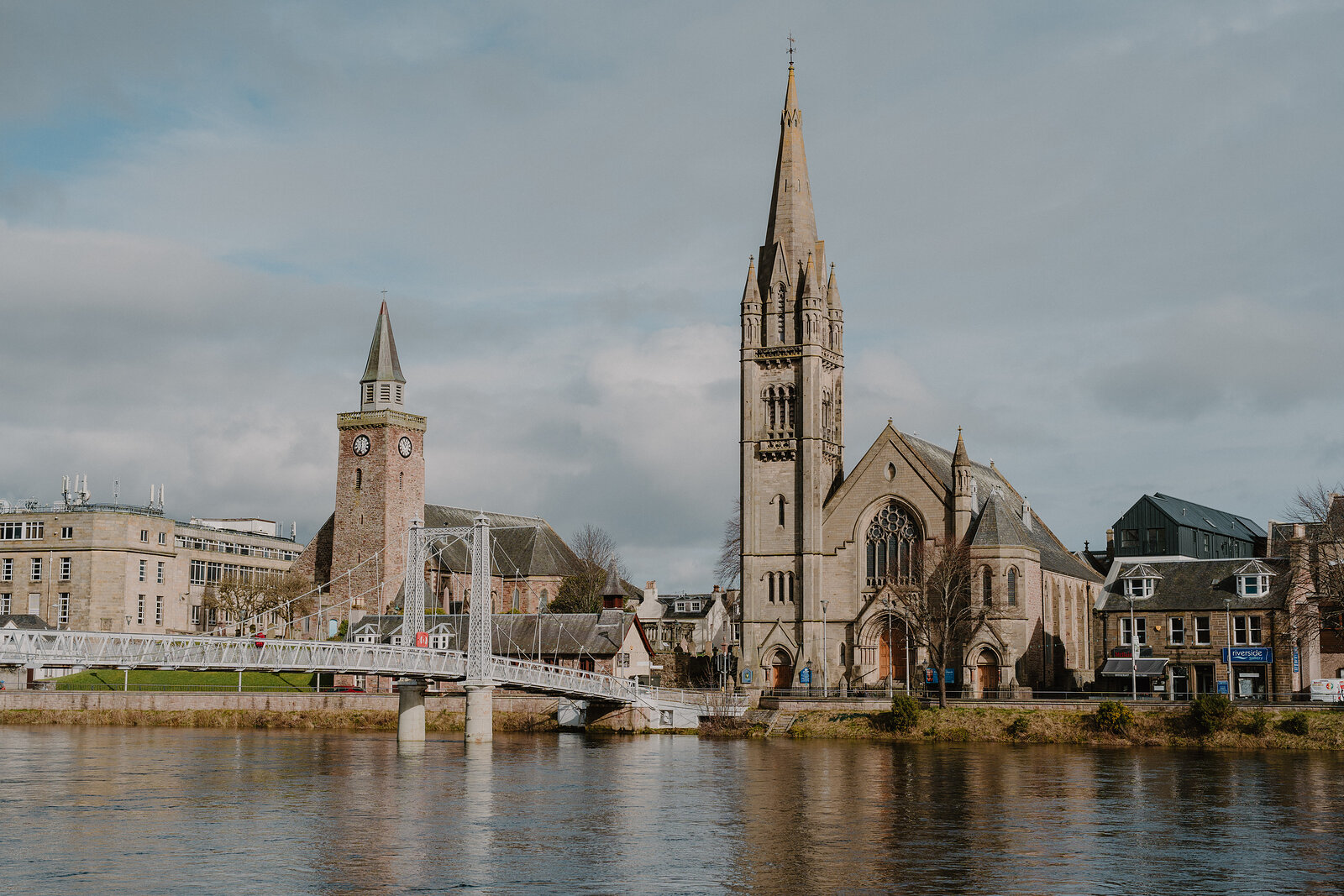 A river runs past a clock tower and a tall spired church, with a white footbridge crossing the water and terraced buildings along the far bank.