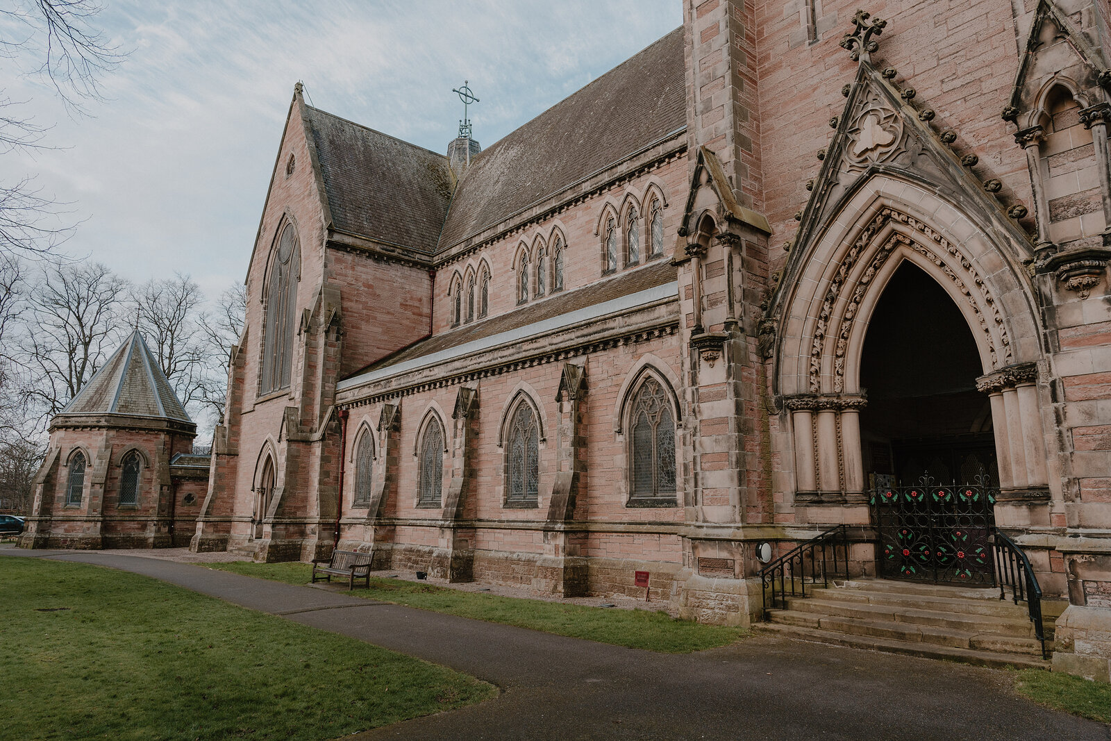 A gothic sandstone church with buttresses and pointed arch windows stands beside a lawn and path, a carved stone porch framing its main door.