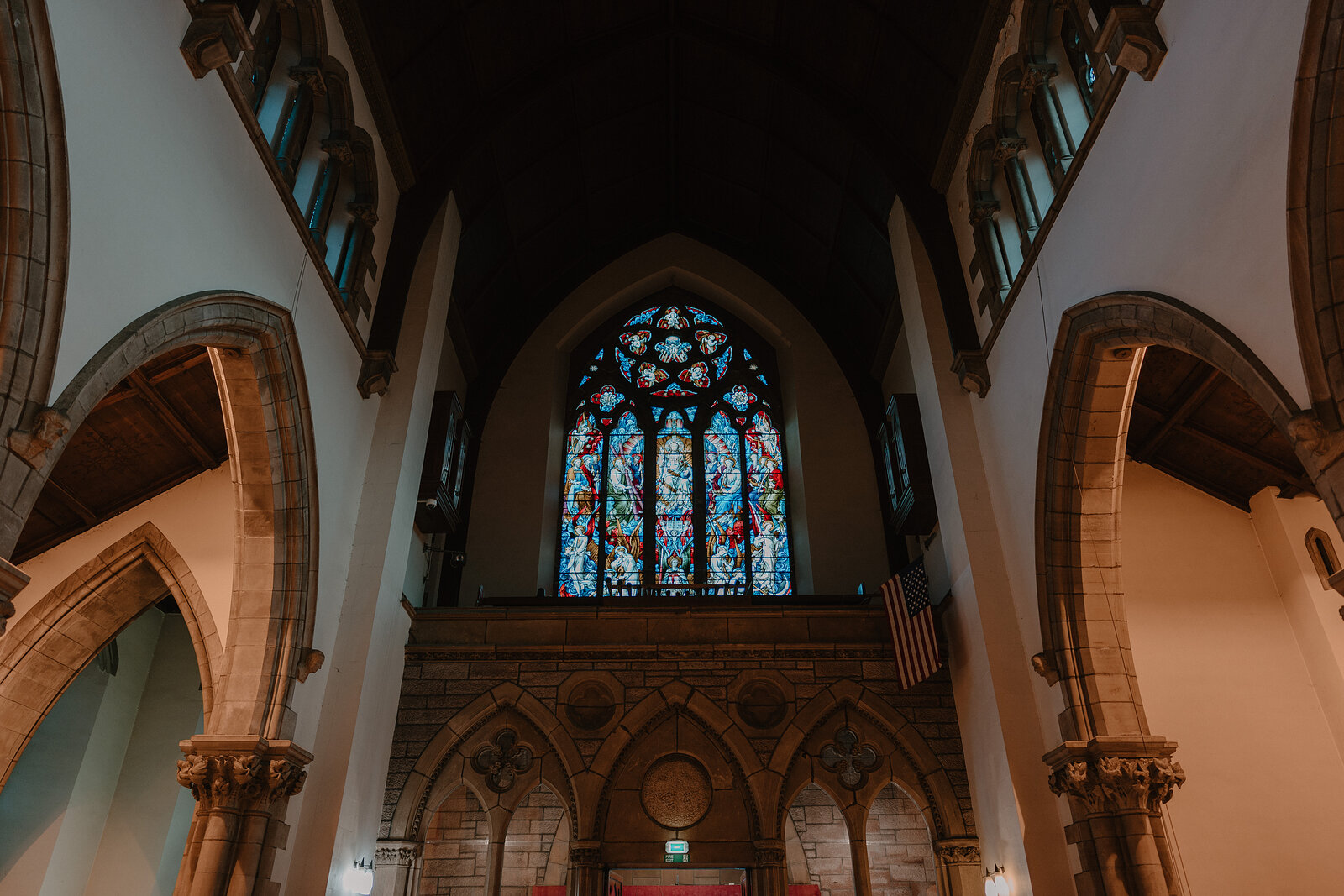 A tall stained-glass window glows above a vaulted stone chancel arch, a small national flag hanging beside carved sandstone columns.
