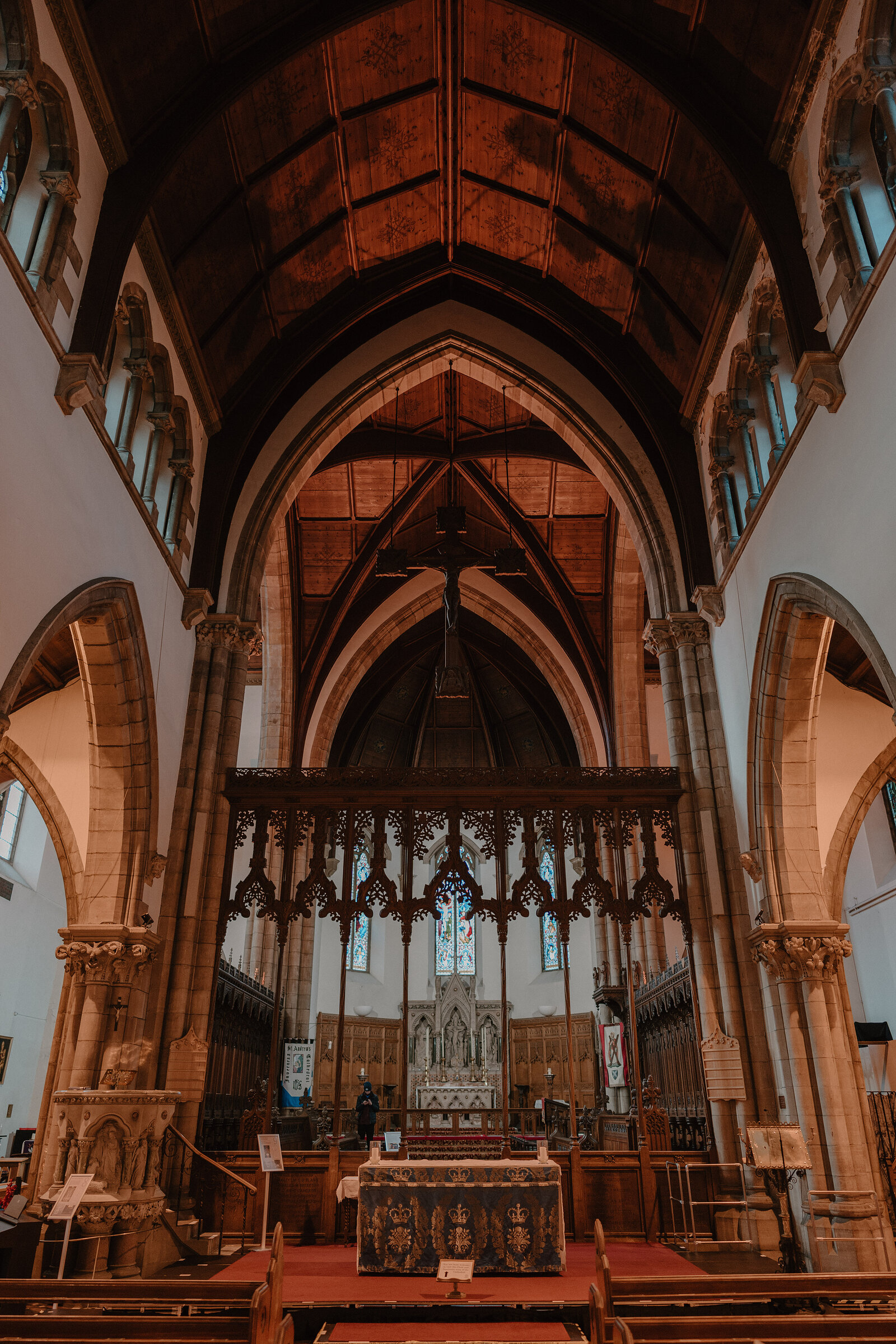 A carved wooden rood screen separates the nave from a chancel with a red-covered altar, beneath a vaulted timber ceiling and sandstone arches.