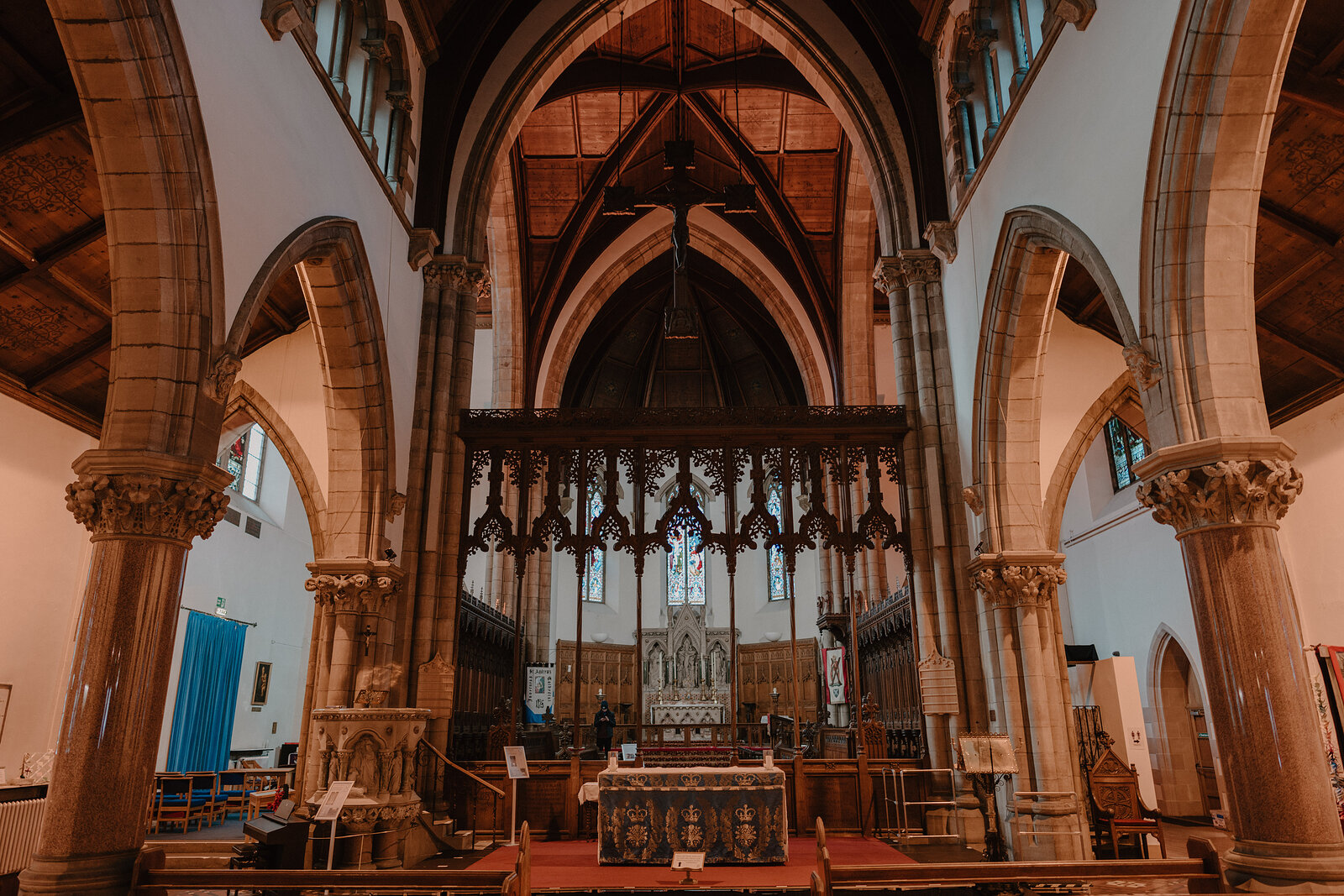 A carved wooden rood screen and a red-draped altar stand beneath a vaulted timber roof, framed by sandstone columns and arched windows.