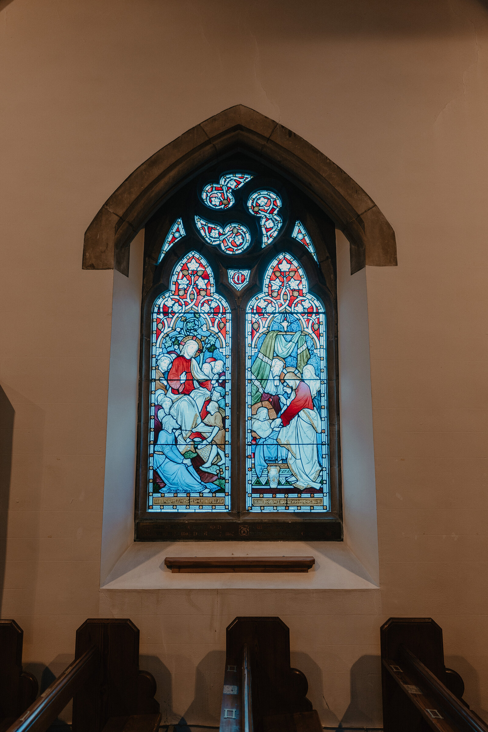 A pointed stained-glass window depicting two robed figures sits within a carved stone arch above wooden pews.
