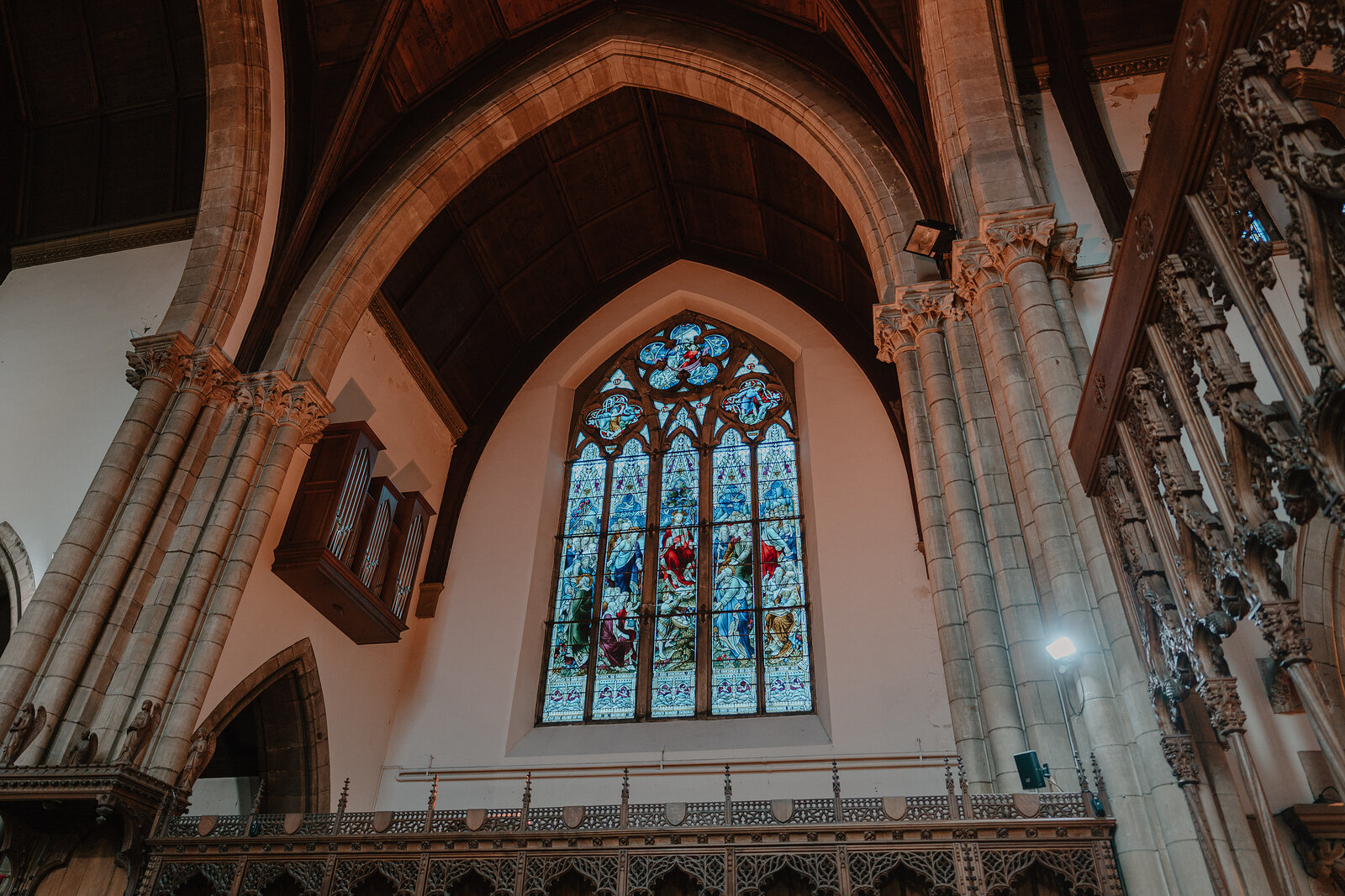 A pointed stained-glass window rises above a carved wooden screen and choir stalls beneath a vaulted stone ceiling.