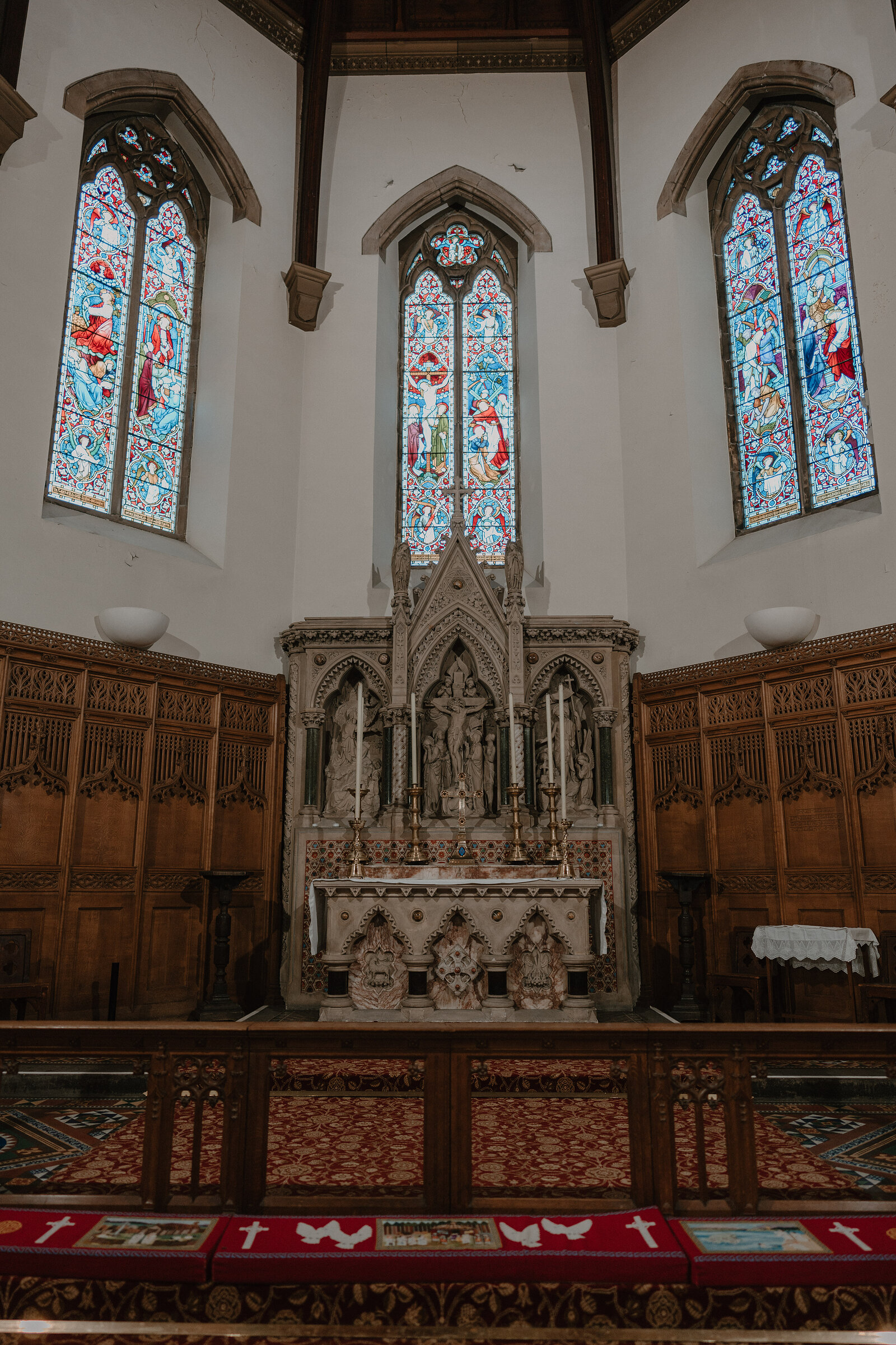 A carved stone altar and reredos stand beneath three tall stained-glass windows, kneelers laid along the altar rail.