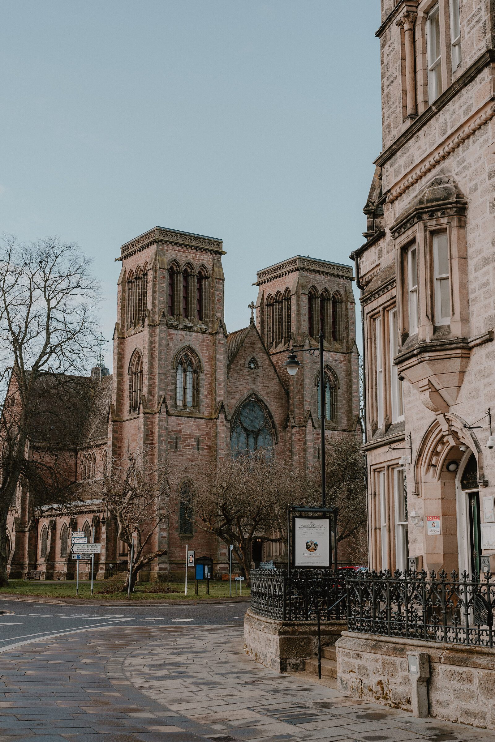 A twin-towered red sandstone cathedral stands on a corner beside a lamp post and railings, bare trees in front of its arched windows.
