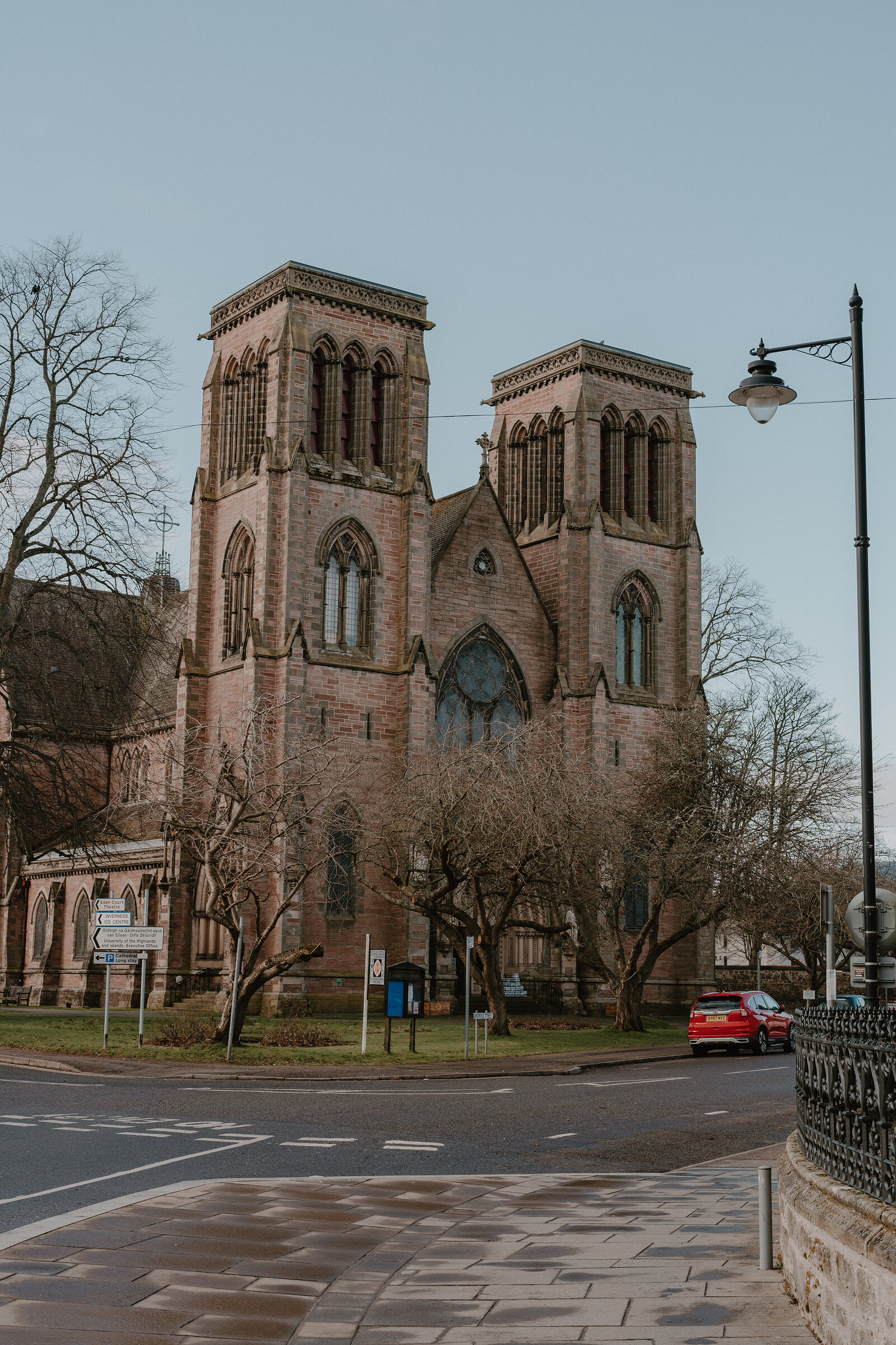 A twin-towered red sandstone cathedral rises above bare trees on a street corner, a parked car and lamp post in front of its railings.