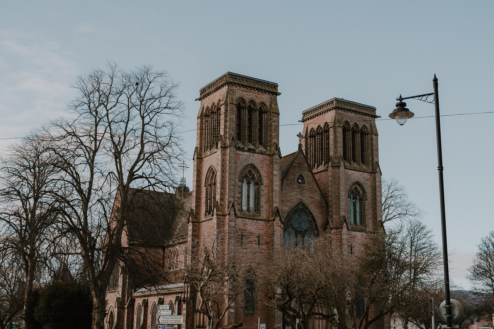 A twin-towered red sandstone church with pointed arch windows stands beside bare trees and a lamp post on a street corner.