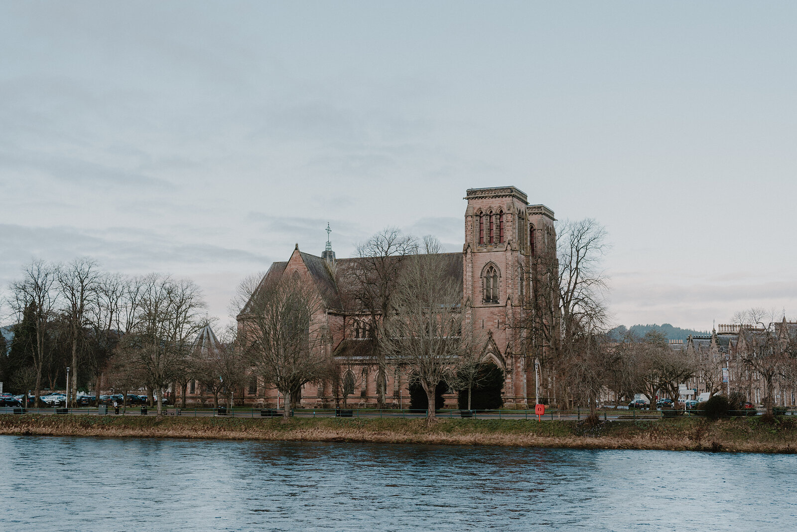 A red sandstone church with a square tower stands beside a river, parked cars and bare trees along the waterfront path.