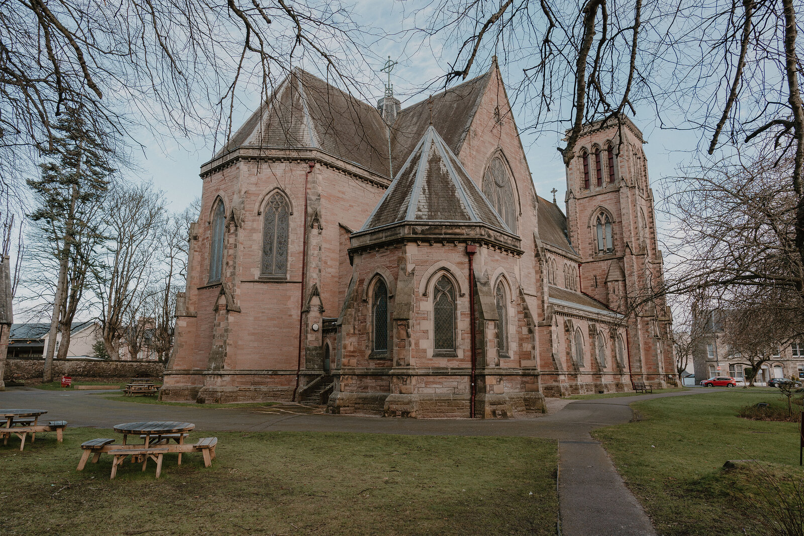 A red sandstone church with a tall tower and steeply pitched gables stands behind a lawn with wooden picnic benches.
