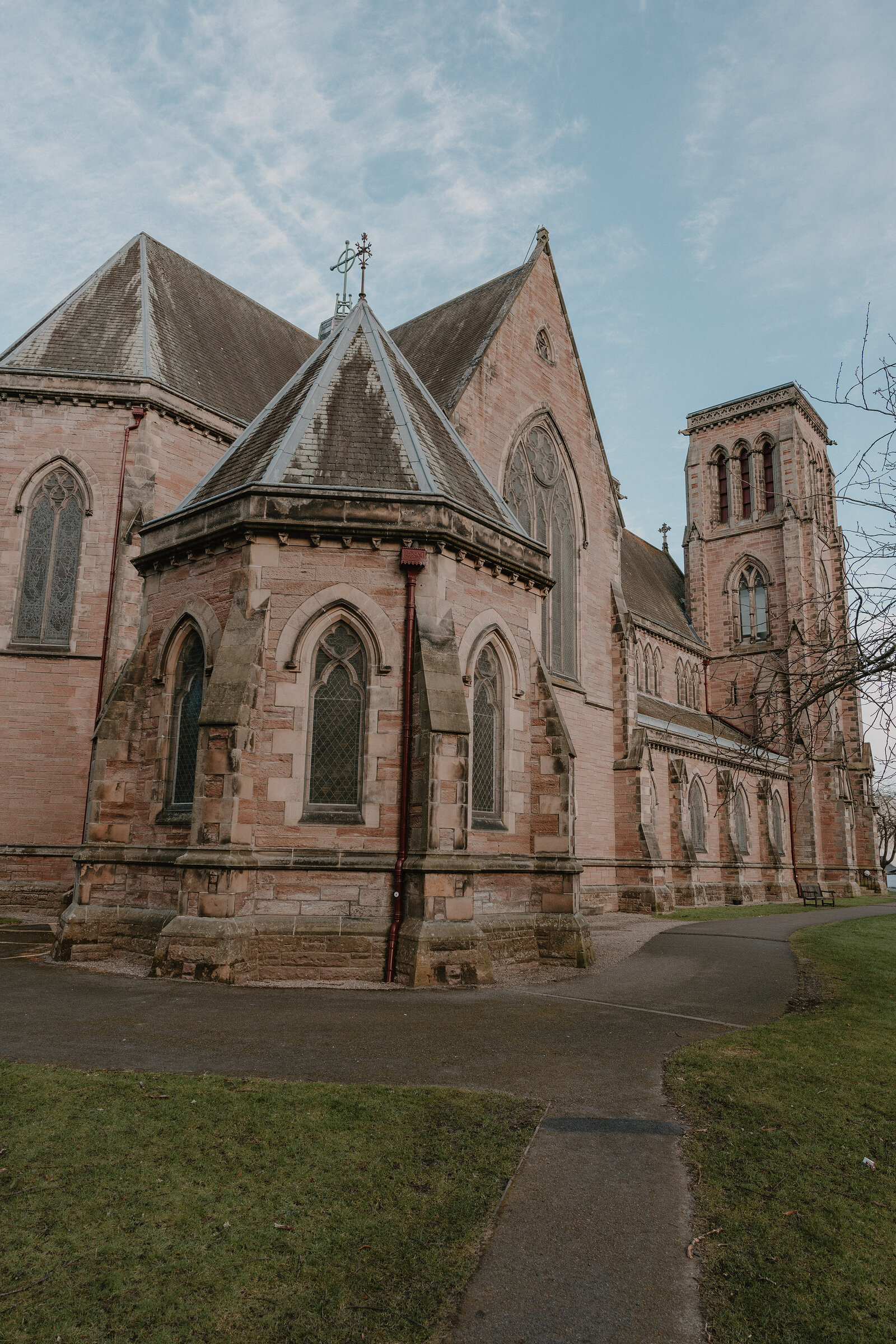 A red sandstone church with an octagonal turret, arched windows and a square tower stands on a corner beside a paved path.