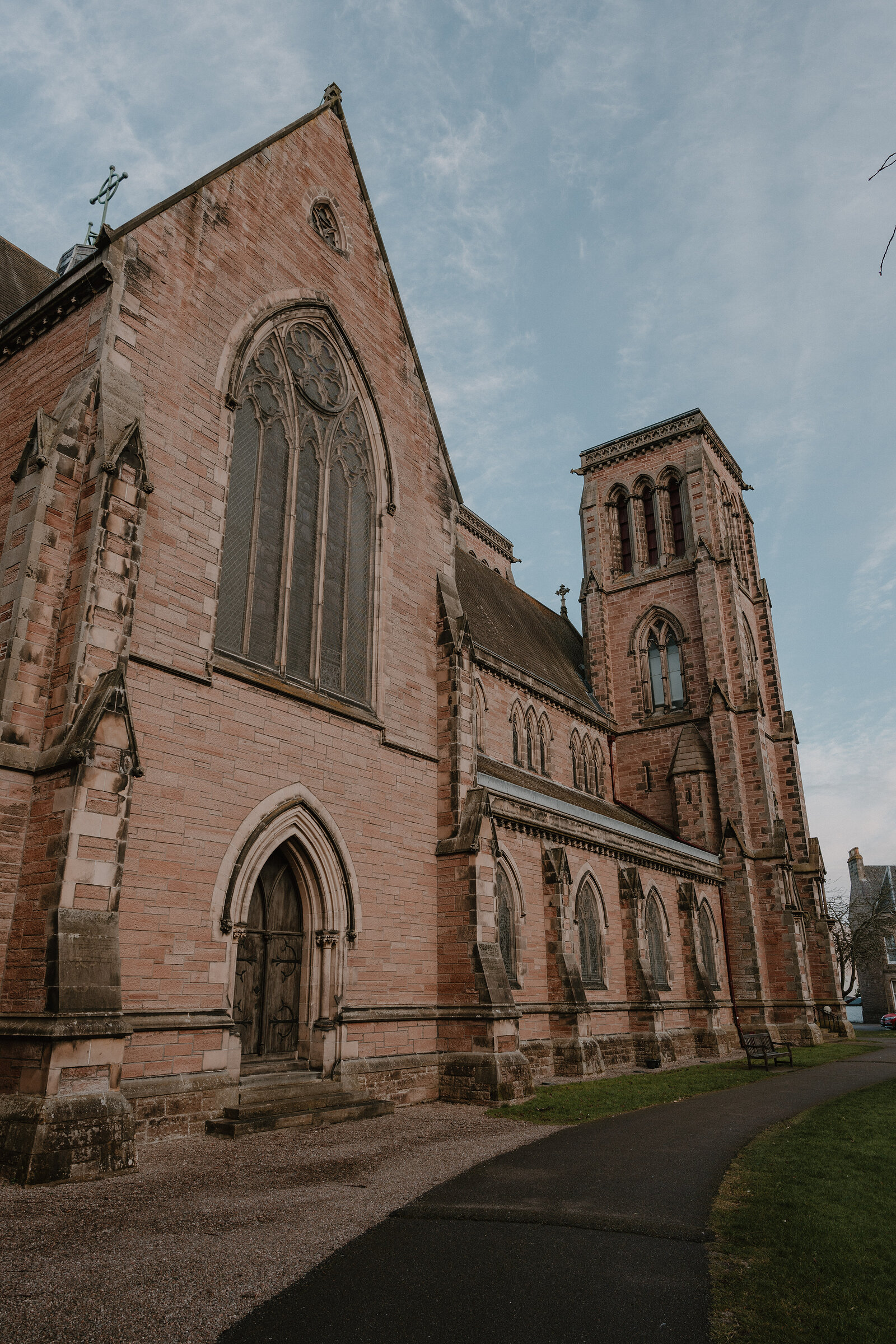 A red sandstone church with a rose window in its gable and a square tower stands beside a paved path and lawn.