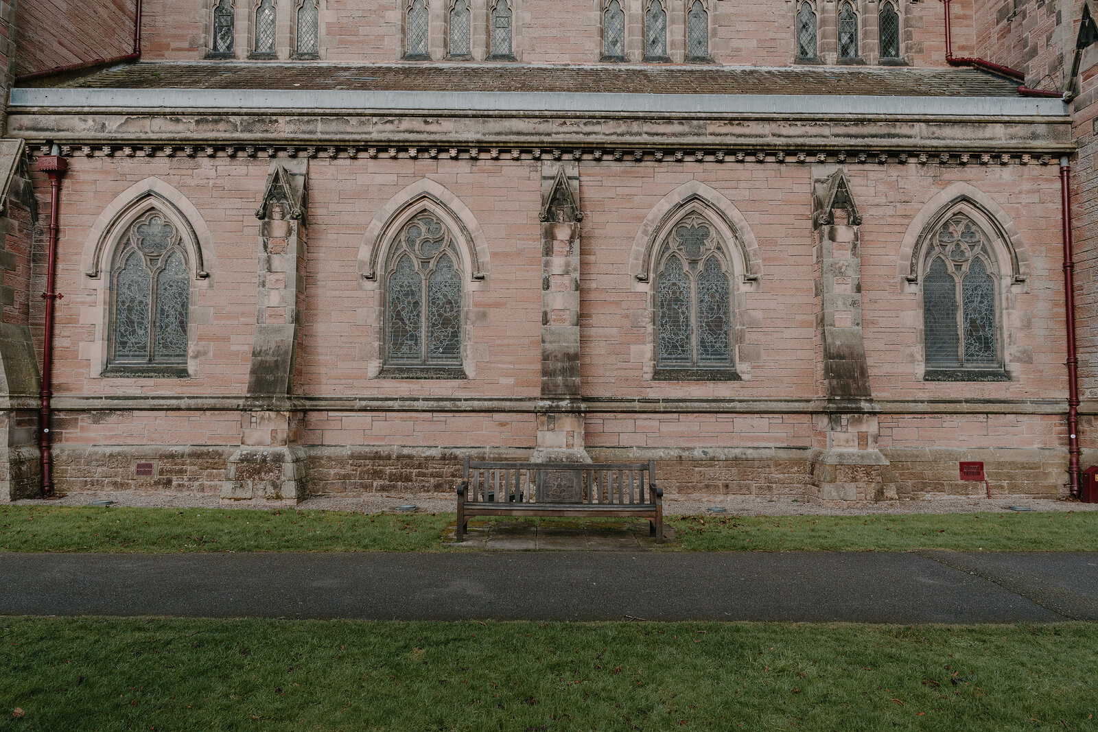 A row of tall lancet windows with stone tracery lines the side of a sandstone church, gargoyles projecting beneath the roofline above a wooden bench on the lawn.
