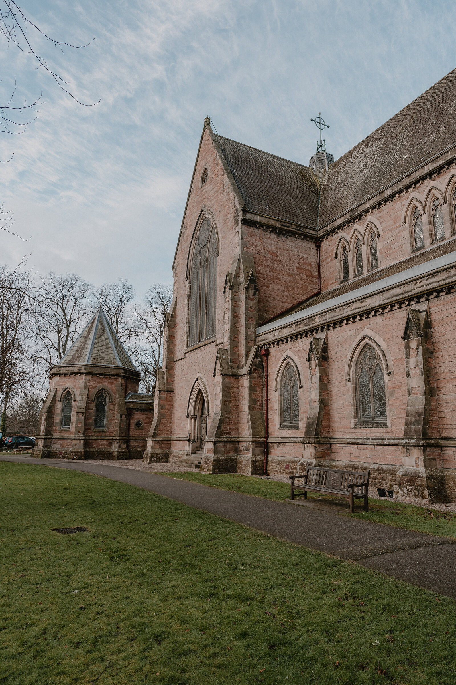 A red sandstone church with tall lancet windows and buttresses stands beside an octagonal turret, a wooden bench on the path in front.