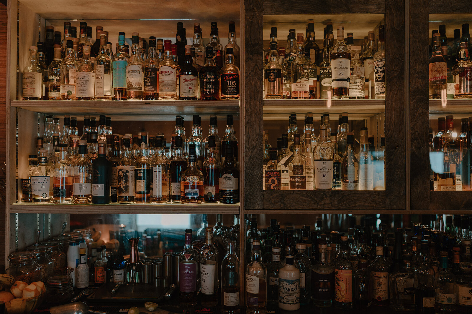 Rows of whisky and spirit bottles fill wooden shelving behind a bar, reflected in a mirrored panel above further bottles on the counter.
