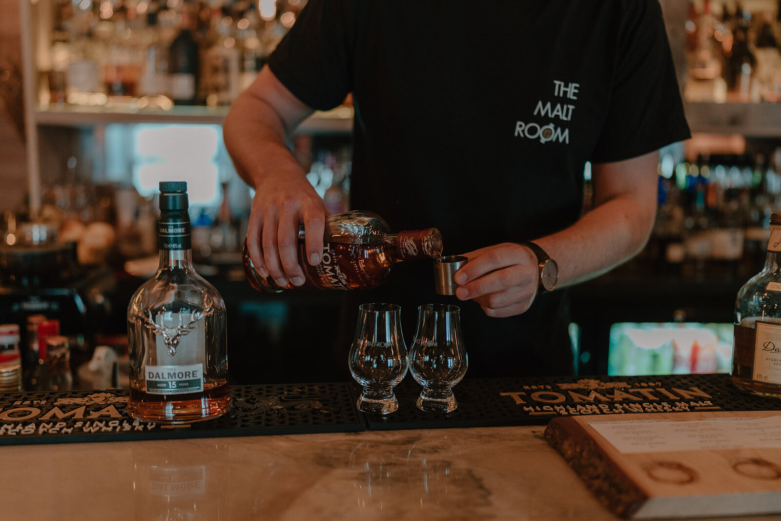 A man in a T-shirt reading "the malt room" pours whisky into two tasting glasses on a bar, bottles from a distillery lined up beside them.