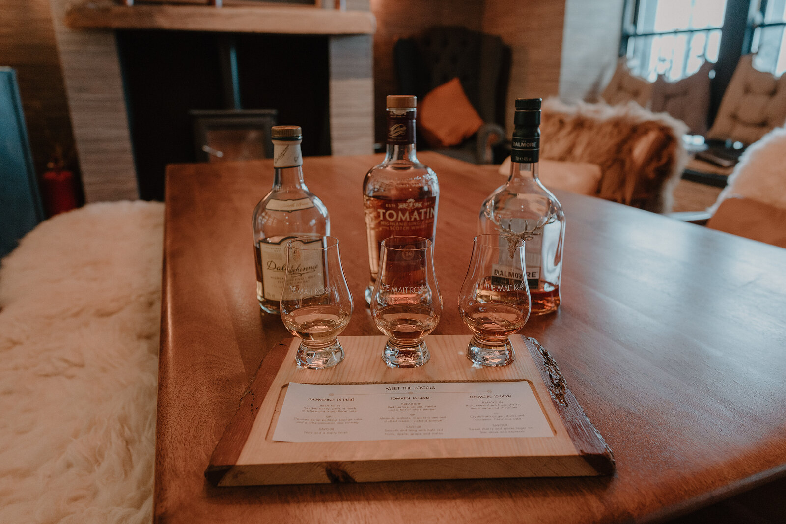Three whisky bottles and tasting glasses stand on a wooden board with a tasting menu beside a stone fireplace and sheepskin-draped armchair.