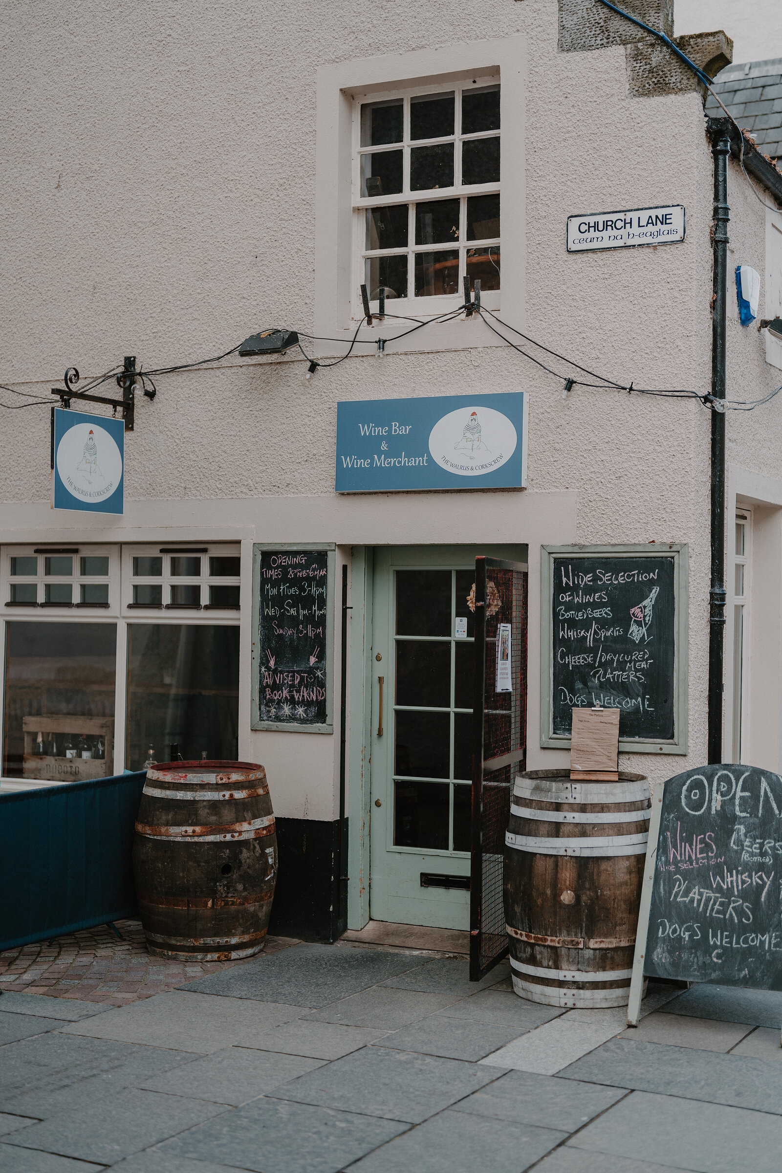 A pale pink building lettered "wine bar, wine merchant" has wooden barrels and chalkboards advertising wine, cheese and whisky flanking its open green door.