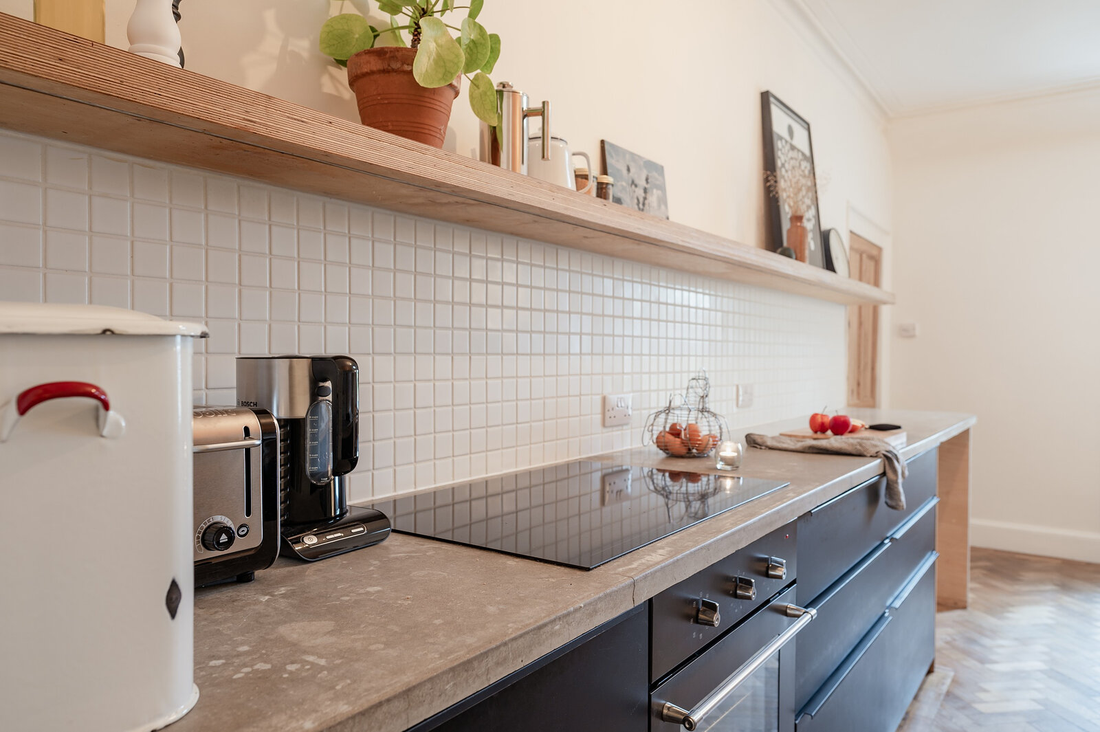 The kitchen's black units and concrete-grey worktop under a shelf of plants.