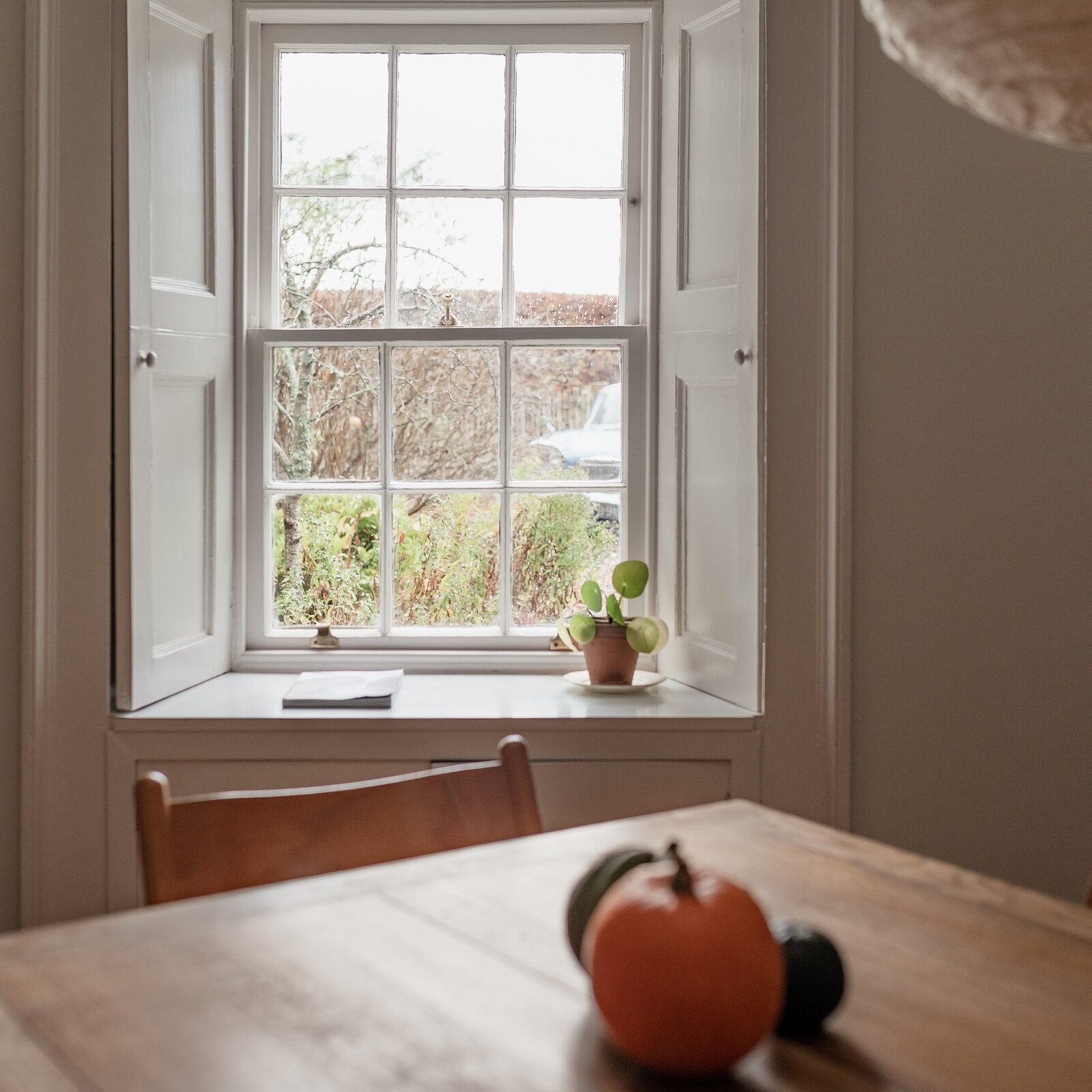 A shuttered sash window above a wooden table with a pumpkin on it, the garden hedge outside.