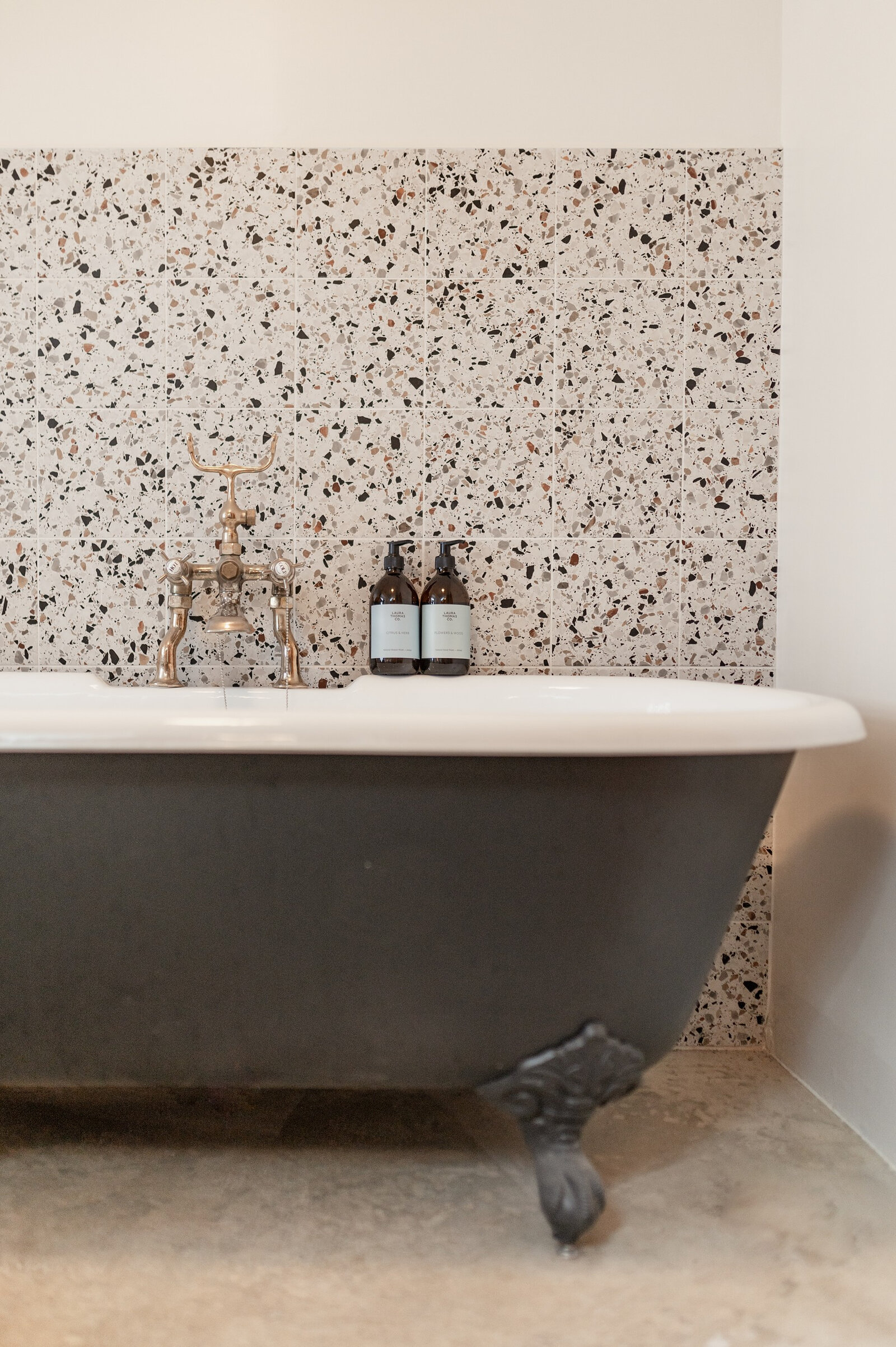 A black claw-foot bath against a speckled terrazzo wall, with brass taps.