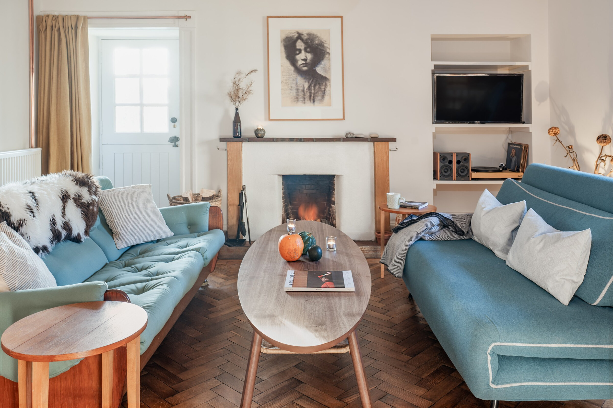 Dell Cottage's sitting room, a fire lit in the open hearth, with teal sofas and sheepskins on an oak parquet floor.