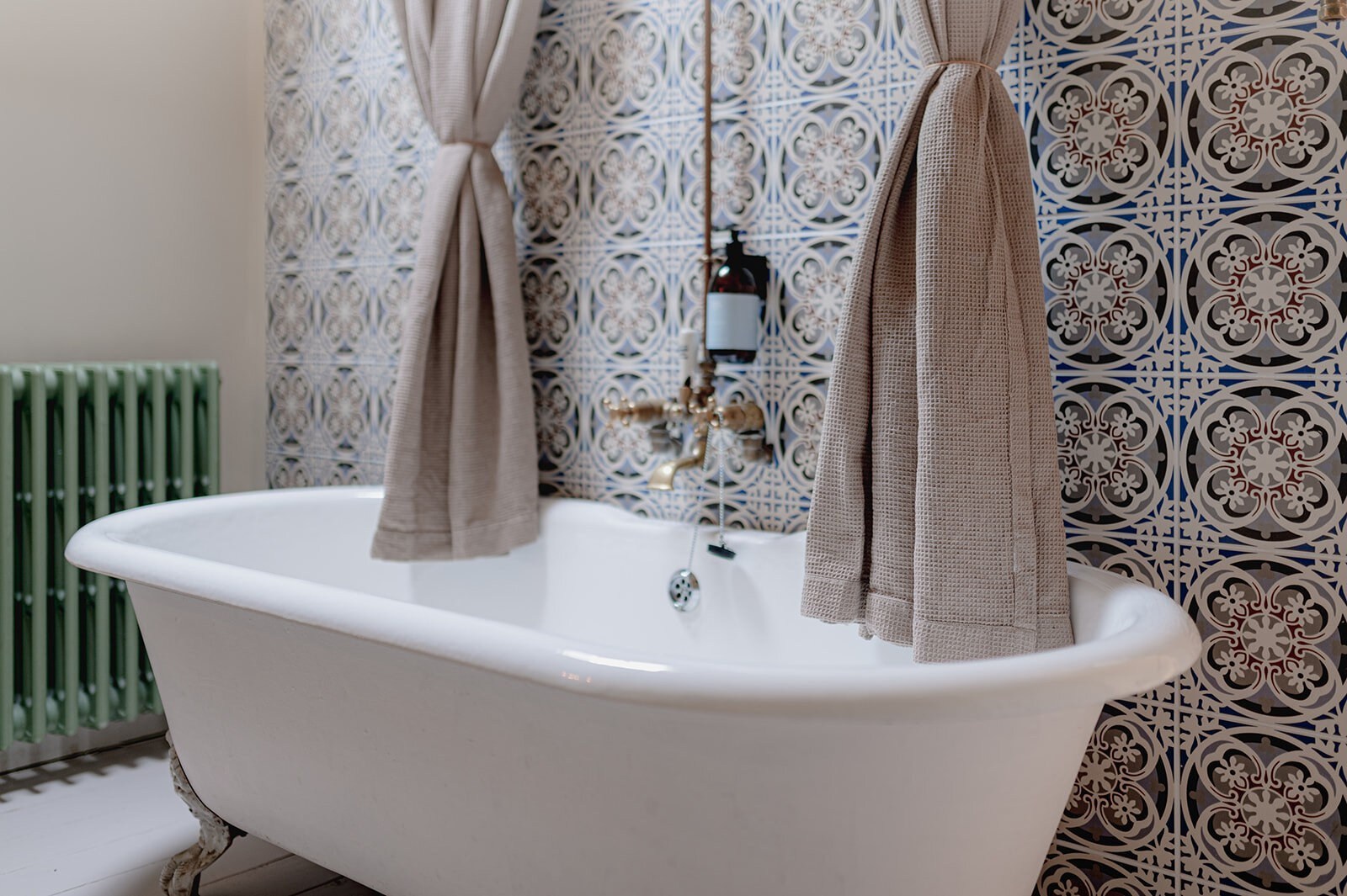 A claw-foot bath against patterned blue and white tiles, with linen curtains and an old radiator.