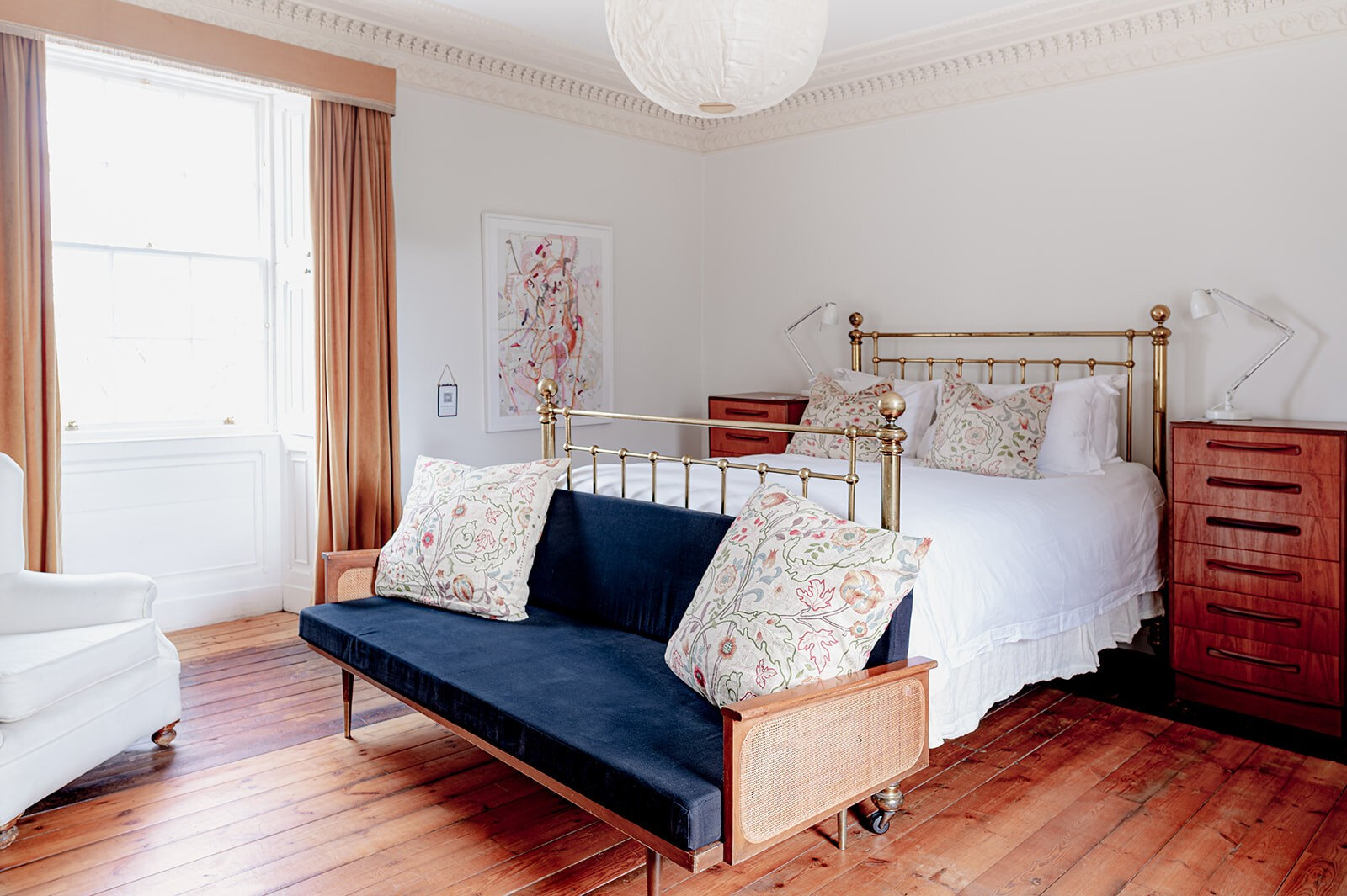 A bright bedroom with a brass bed, a navy daybed at its foot and tall sash windows.