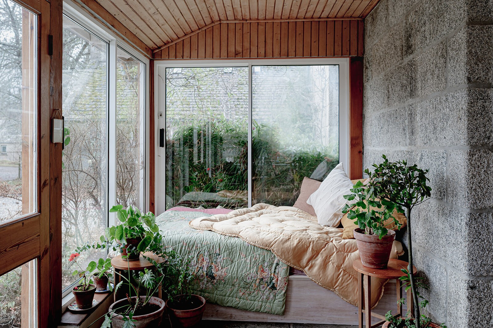 The sun porch, its glass walls looking onto the garden, with a day bed, cushions and pots of plants.