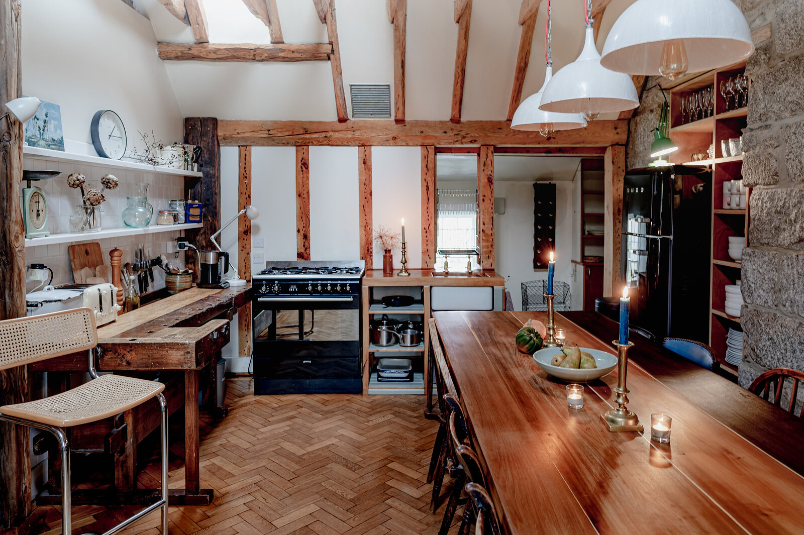 The farmhouse kitchen under old timber beams, with a long table set with candles, a range cooker and open shelves.