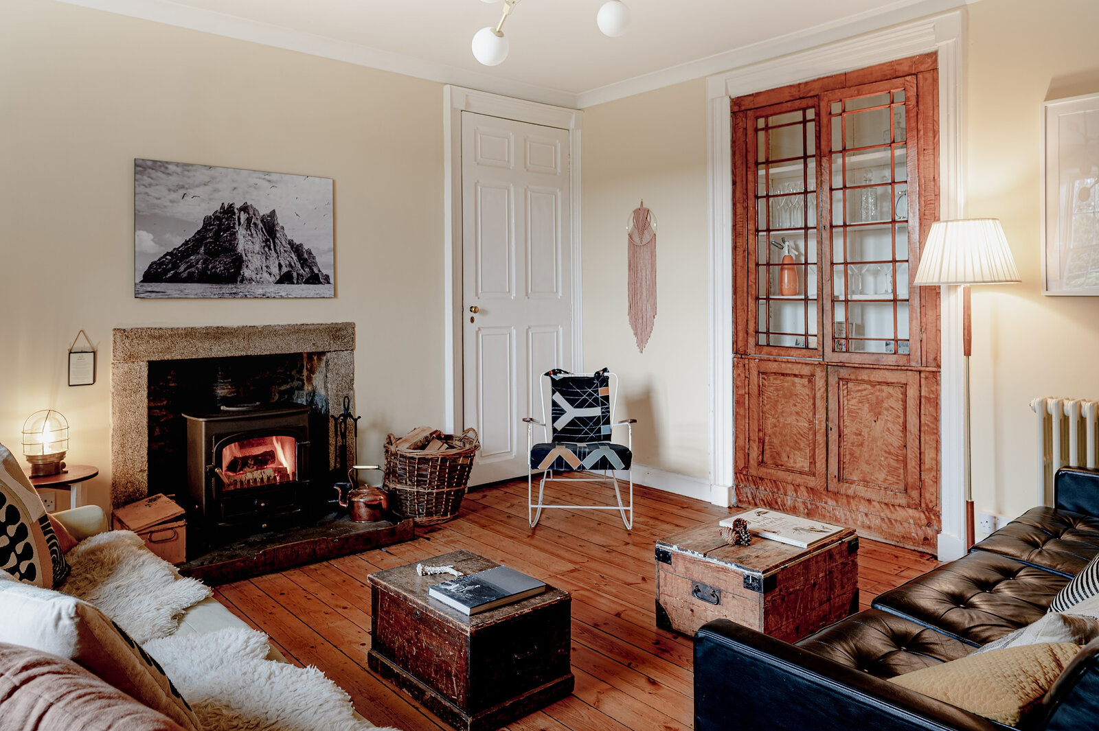 The sitting room with a fire lit in the stove, leather sofas, a tall glazed bookcase and a painting of a mountain.
