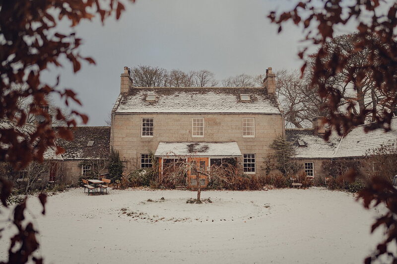 Dell Lodge under snow, its pale stone front and dormers seen through red autumn leaves across the white lawn.