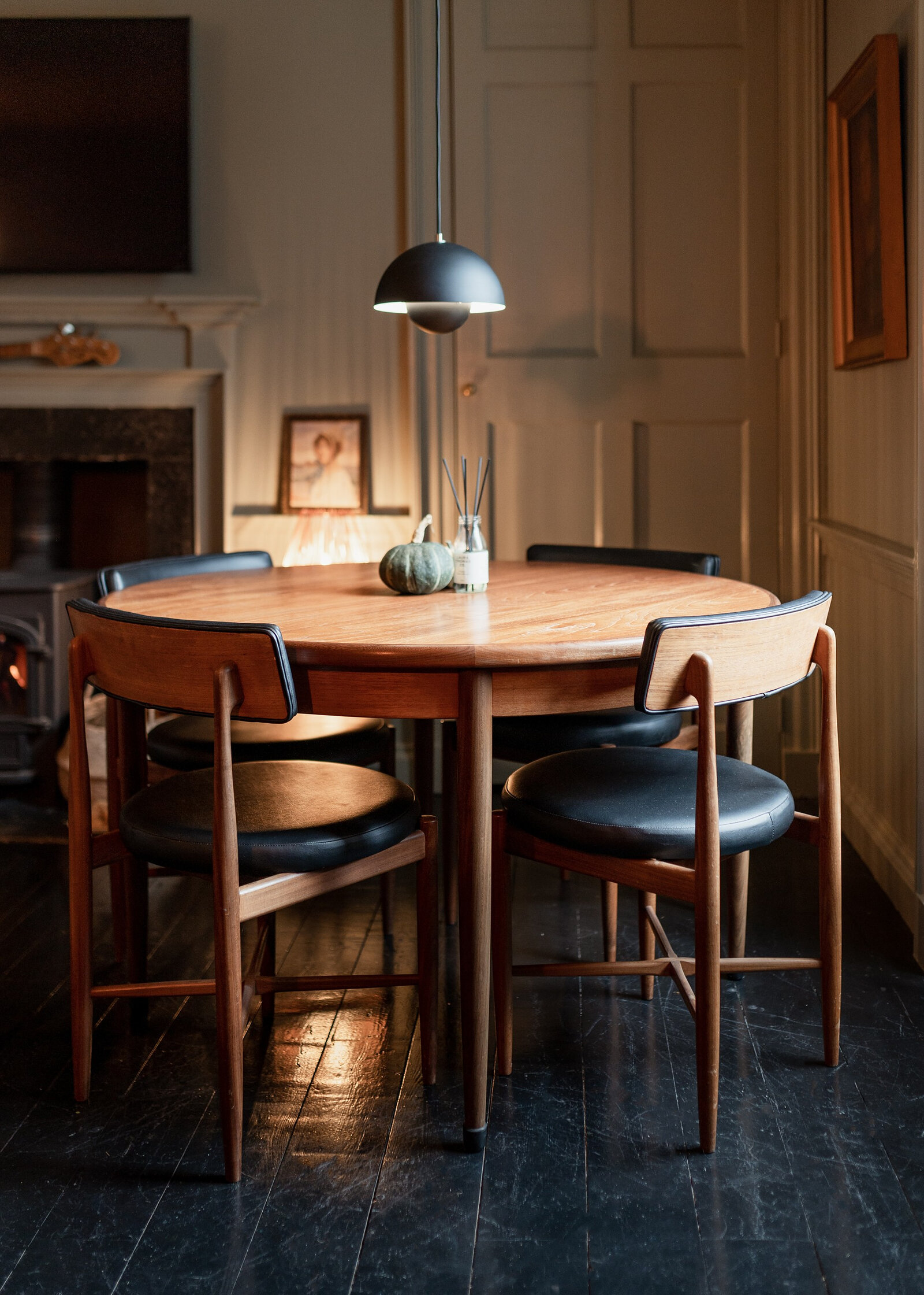 A round wooden table and dark leather chairs under a pendant lamp in a panelled dining room.