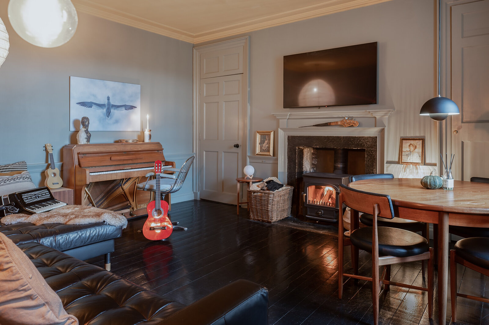 A second sitting room with a piano, a guitar, an open fire and a round dining table under a hanging lamp.