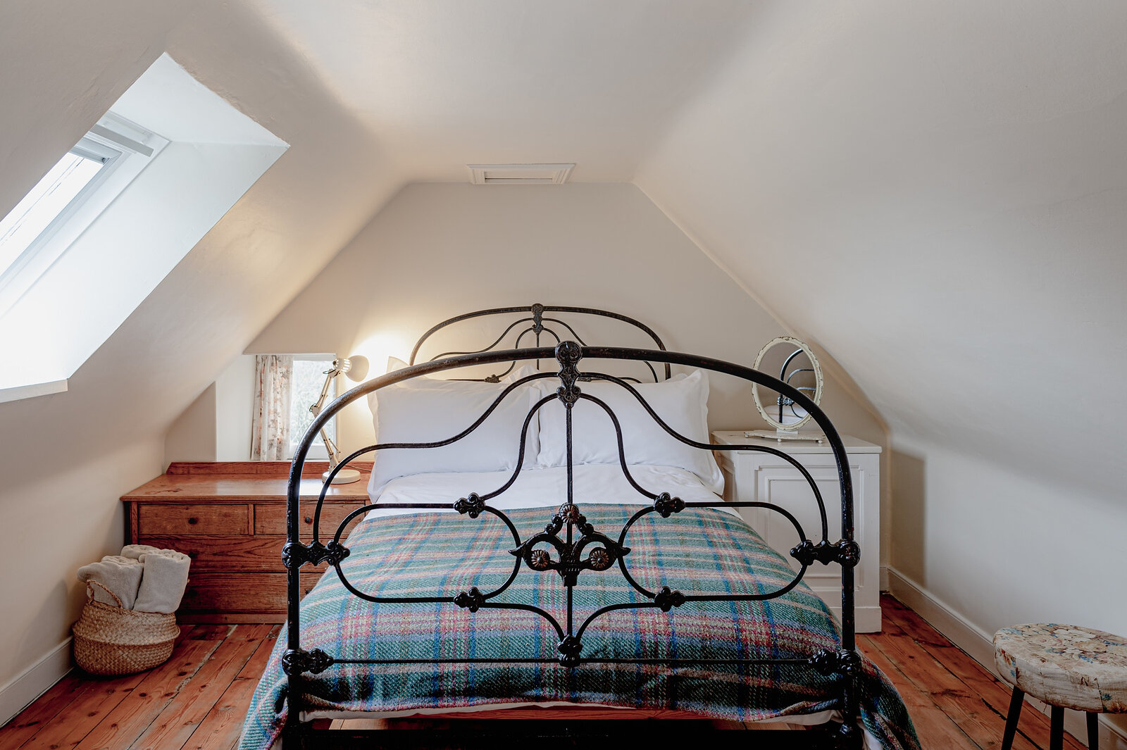An attic bedroom with an iron bedstead, a tartan blanket and a skylight in the sloping ceiling.