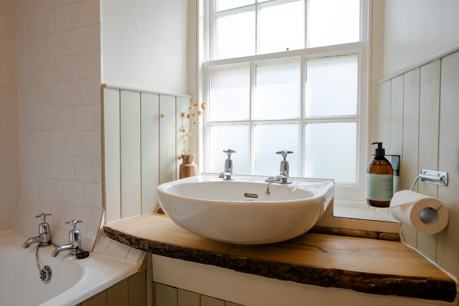 A basin on a slab of live-edge wood beneath a sash window, with the bath alongside.