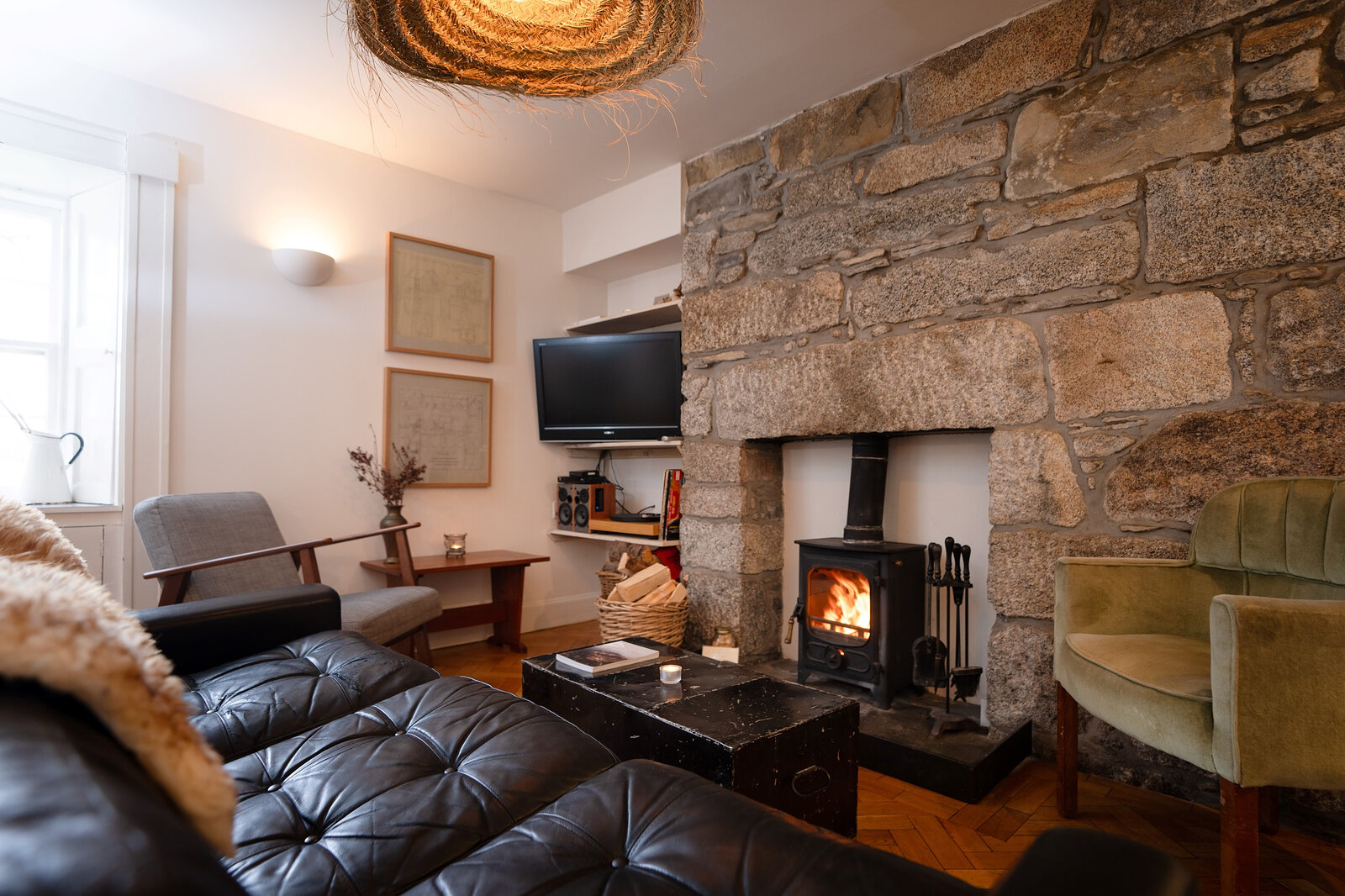 The sitting room, a stove lit in a rough granite fireplace, with a leather sofa, a green armchair and a sheepskin.