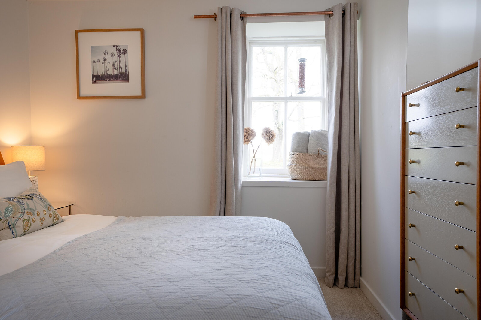 The double bedroom in soft light, with a grey quilt, a chest of drawers and a window with long curtains.