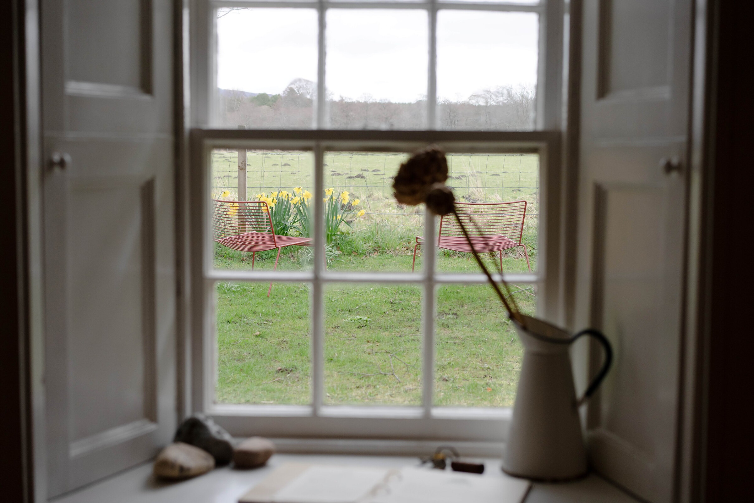 A shuttered window looking onto the garden, its lawn, garden chairs and daffodils, with dried flowers in a jug on the sill.