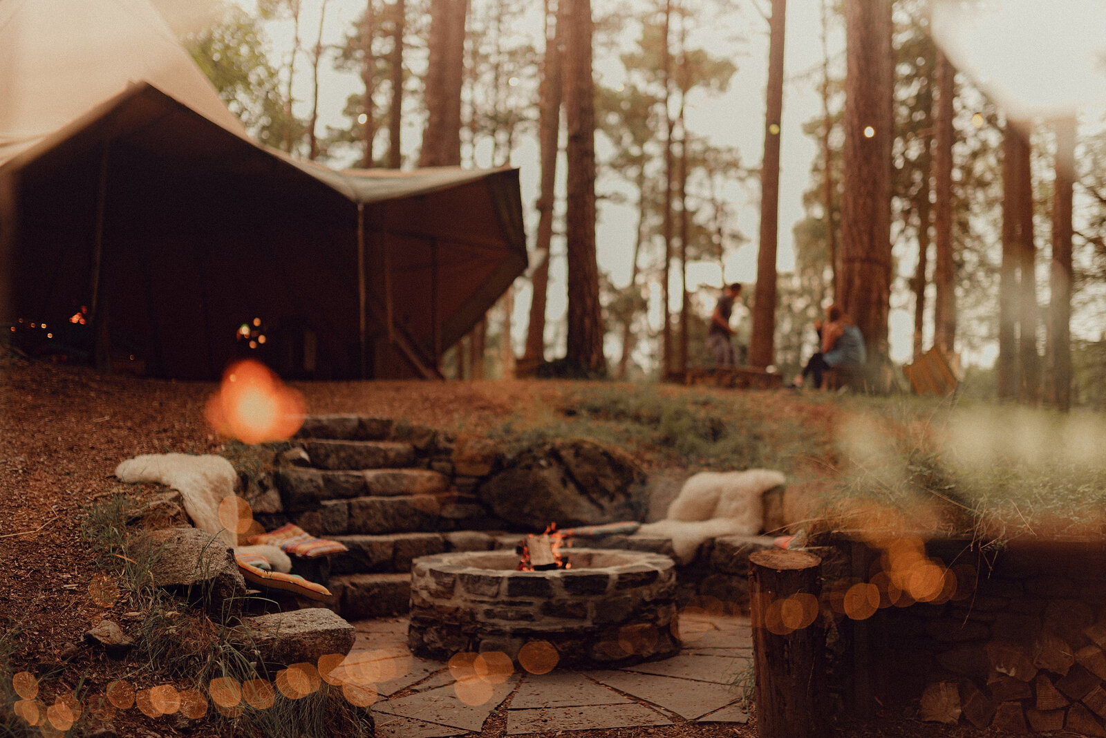 The stone fire pit beside the canvas tipi, a fire lit in it, with sheepskins on the stone seats and the pines behind.