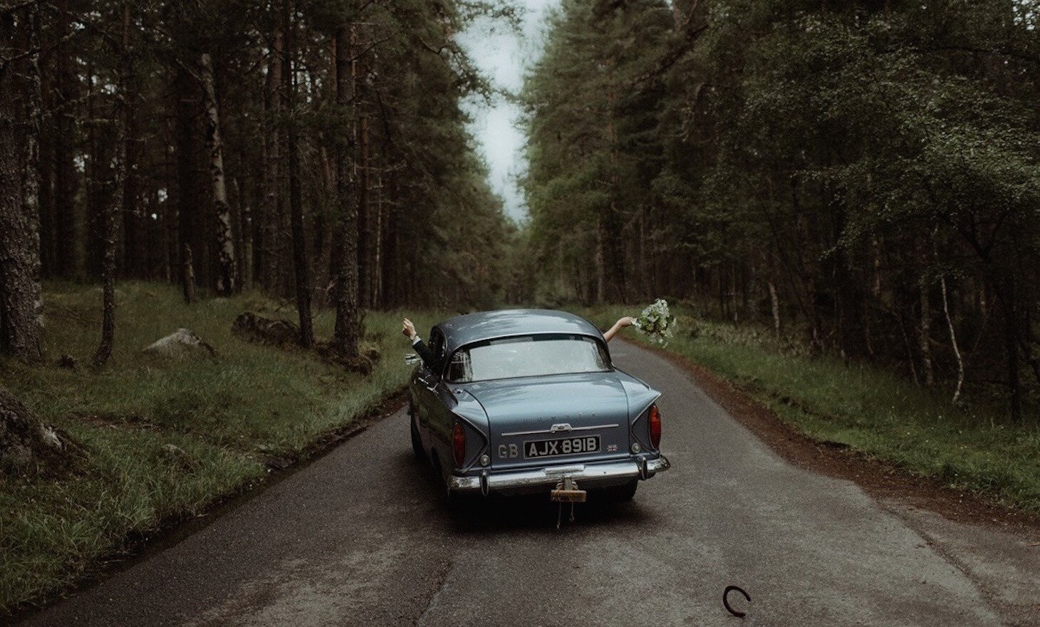 A classic blue wedding car driving away down a road through the pine forest.