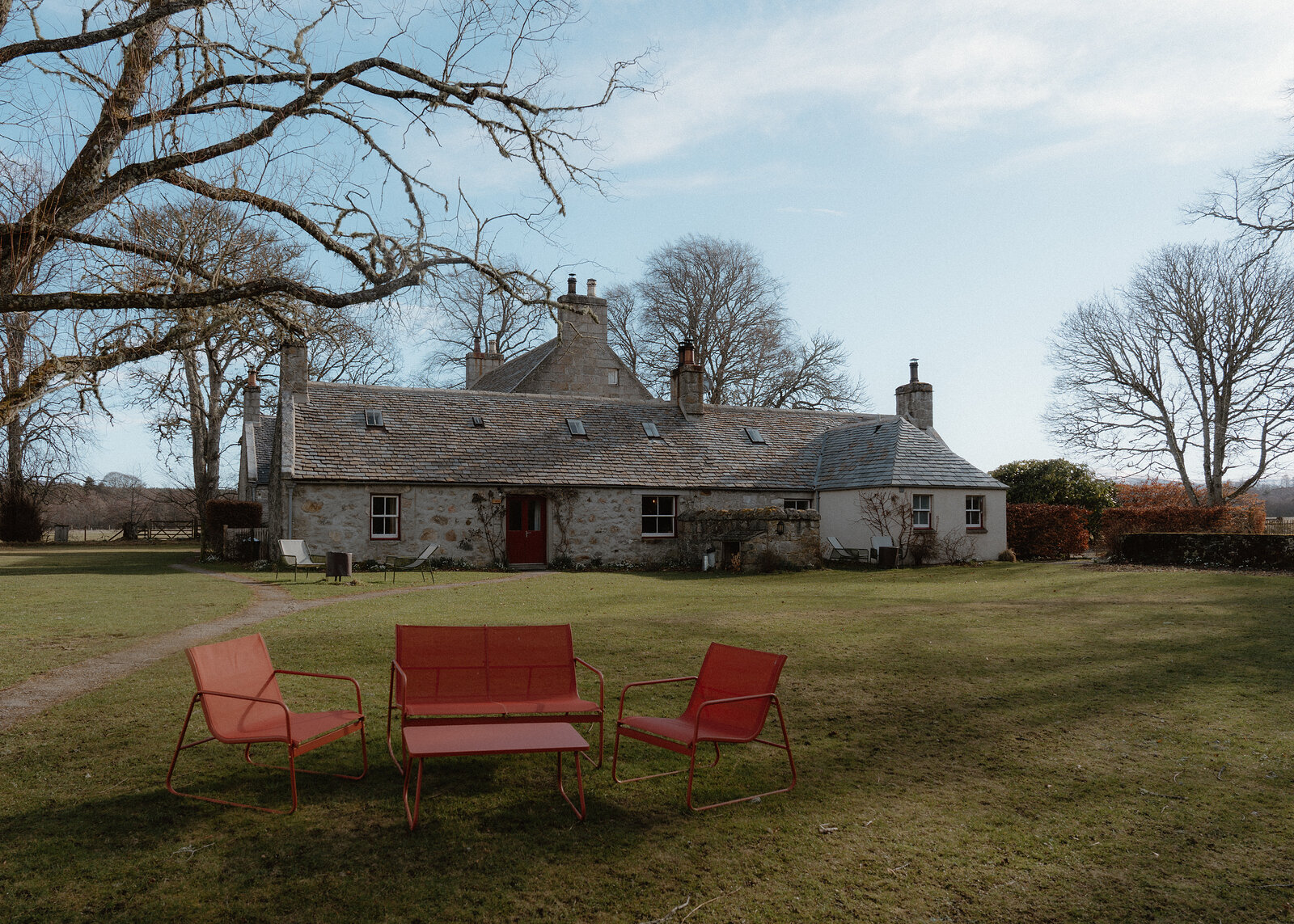 The Dell across the lawn in winter sun, its long stone range and slate roofs under bare trees, three red garden chairs in front.