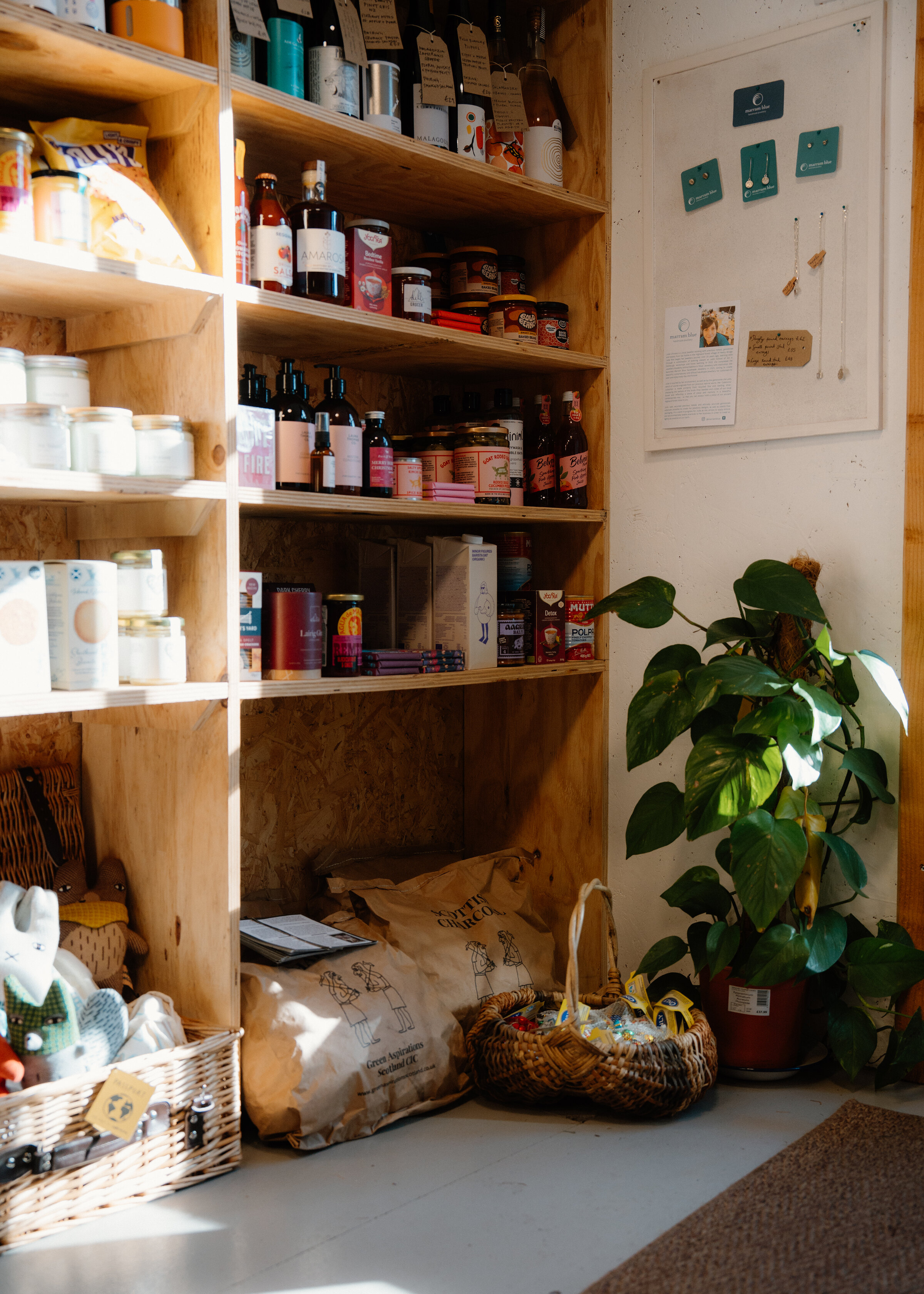 A sunny corner of the Dell Grocer, with shelves of bottles and jars, a cheese plant and baskets on the floor.