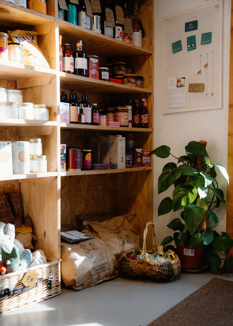 A sunny corner of the Dell Grocer, with shelves of bottles and jars, a cheese plant and baskets on the floor.